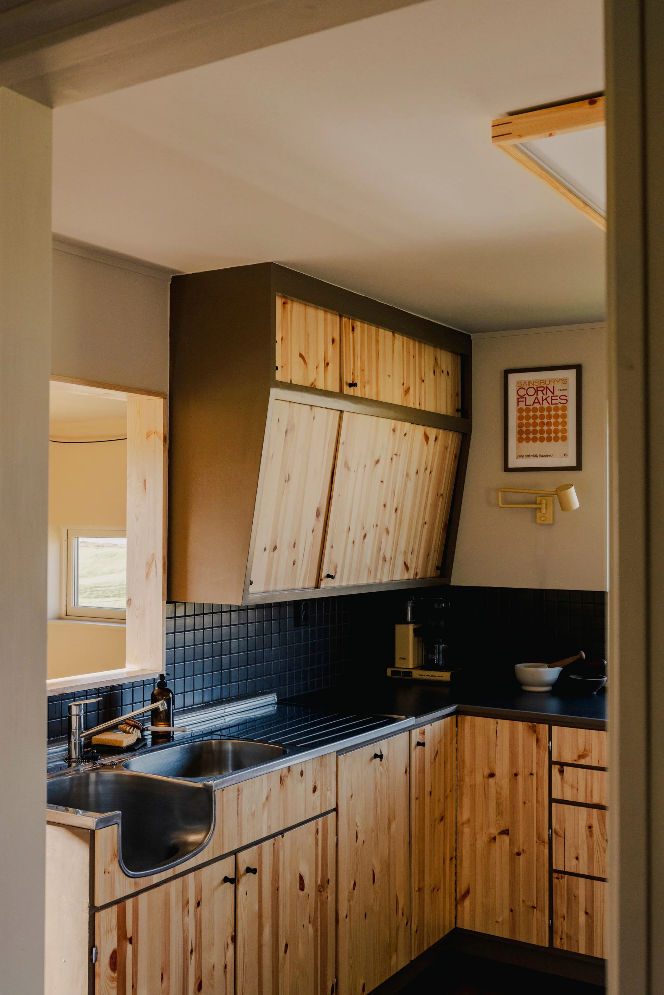 A kitchen with wooden cabinets, a black countertop, a stainless steel sink, a window, and framed artwork on the wall.