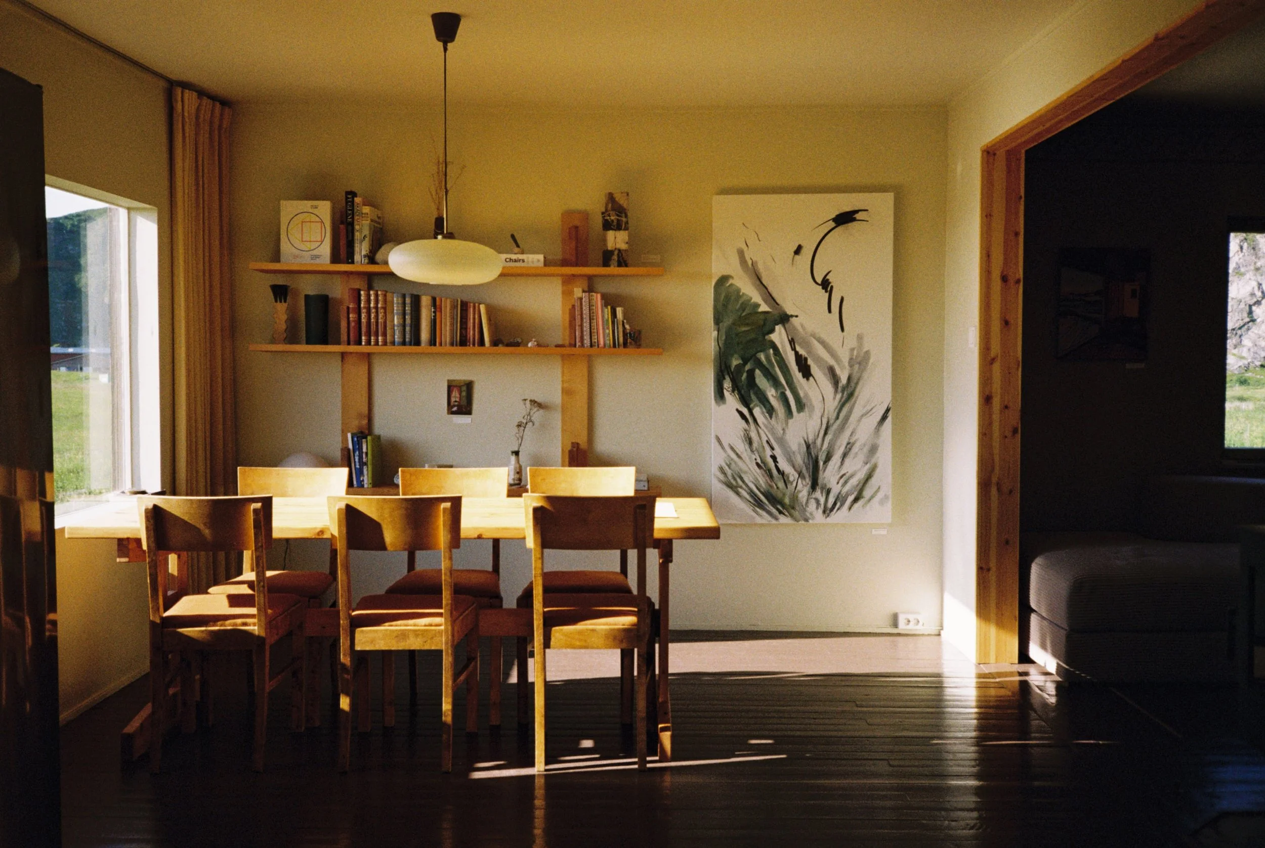 A wooden dining table with seven matching chairs in a well-lit room with large windows. A floating shelf holds books and decorative items, and an abstract painting adorns the wall. The room features natural wood accents and warm lighting.