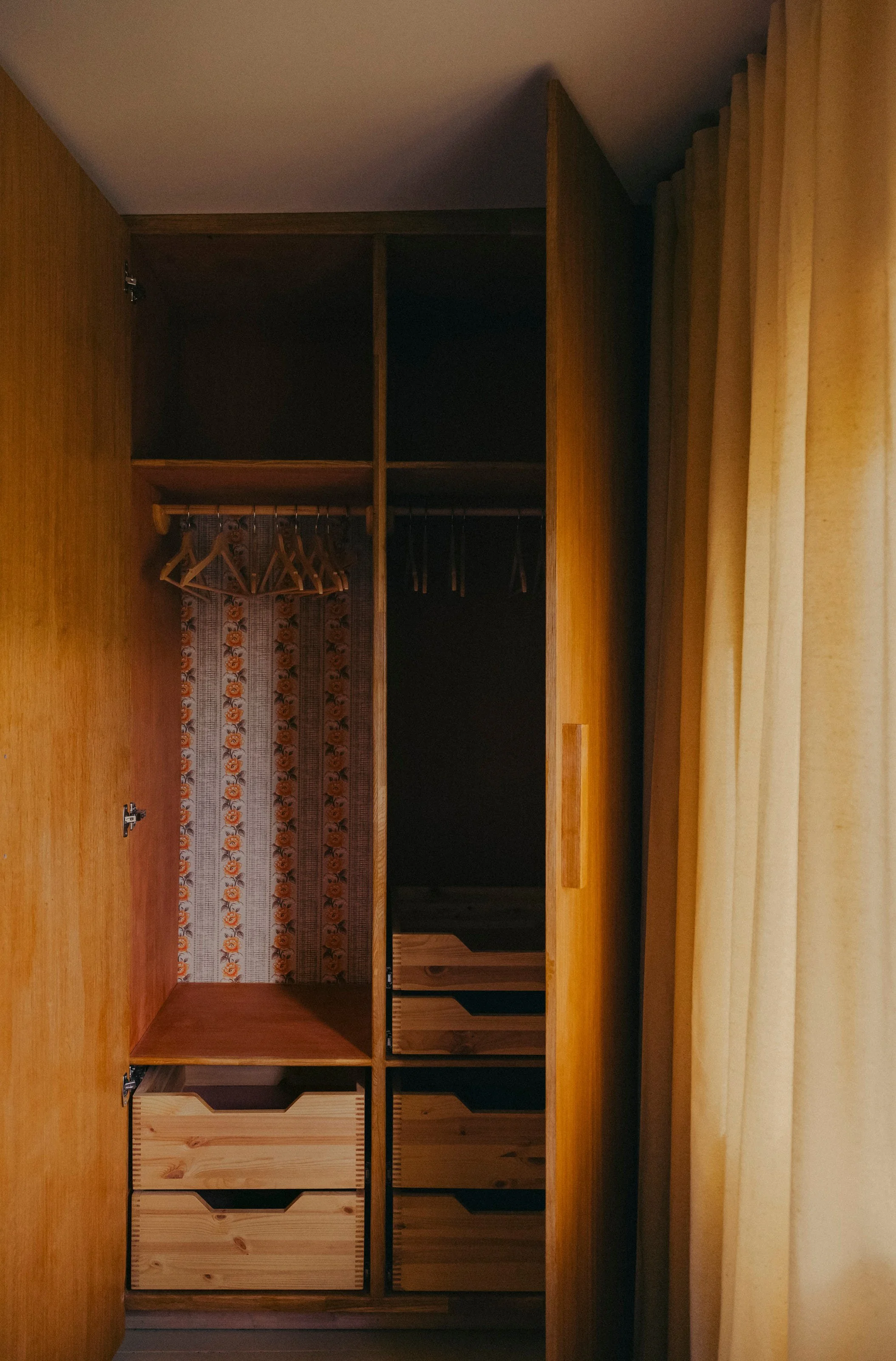 Empty wooden wardrobe with shelves and hanging space, located next to a window with yellow curtains.