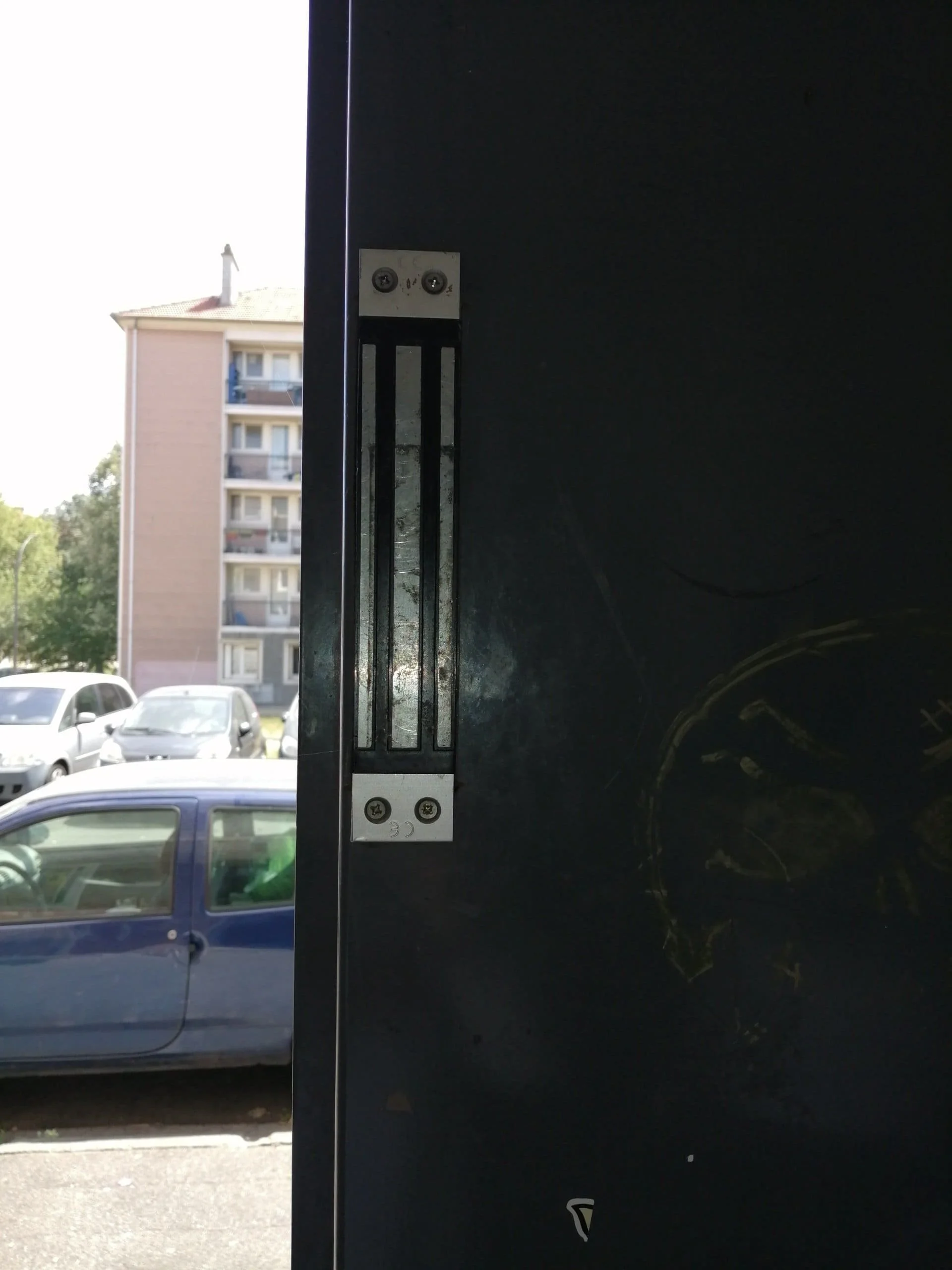 Un loquet de porte en gros plan avec un bâtiment résidentiel flou en arrière-plan, et plusieurs voitures garées à proximité.