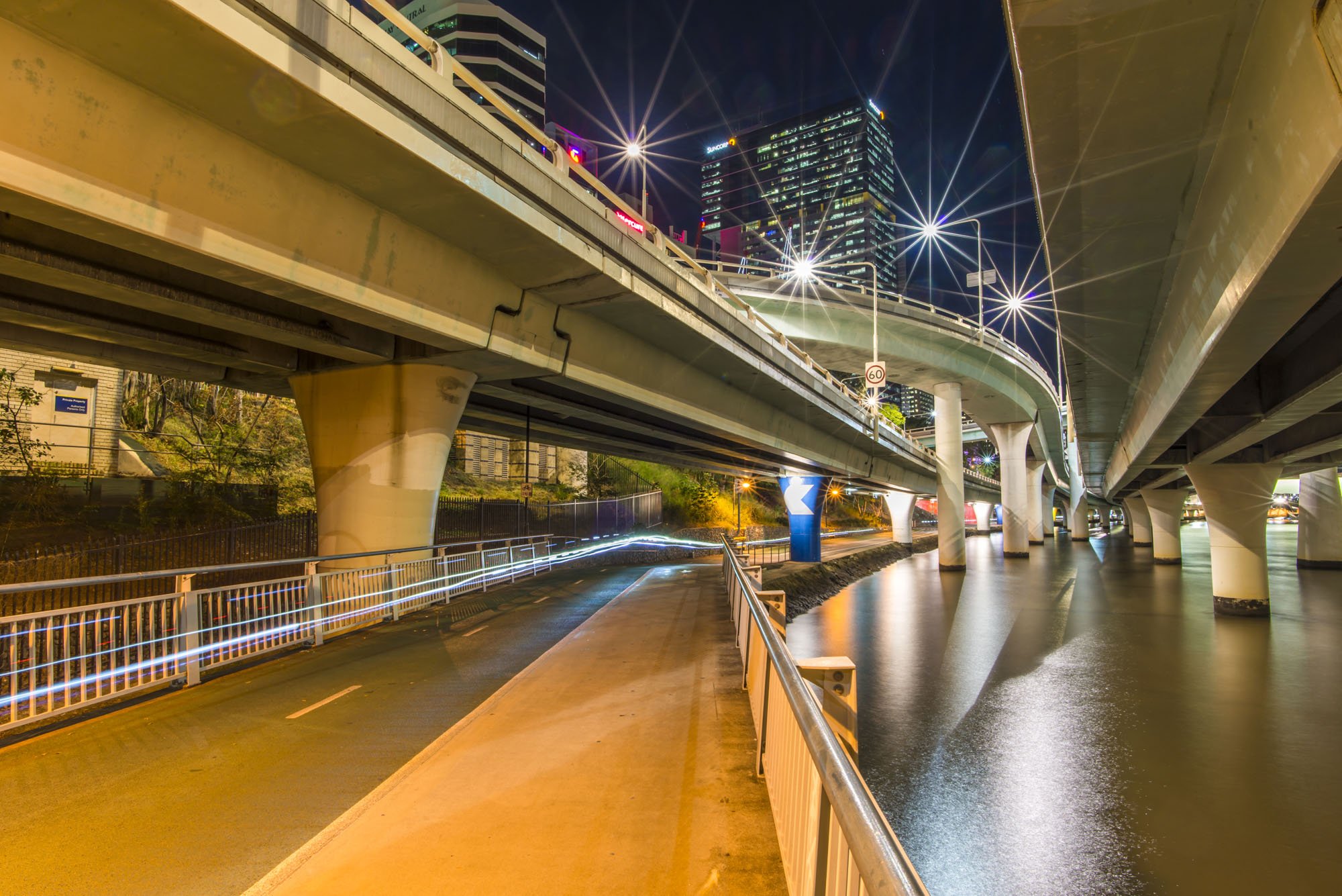 A low angle photo of the underneath of roads and intersections taken from a cycle lane in Brisbane, Australia. It's a night scene with a dark sky and bright street lights that have a starburst effect. The roads spiral together and lead the eye toward