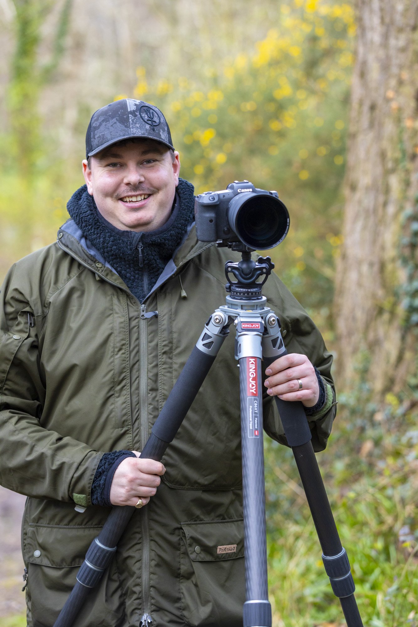Professional nature and landscapes photographer Jack Lodge poses next to his camera on a tripod and smiles for a headshot in an outdoors setting.