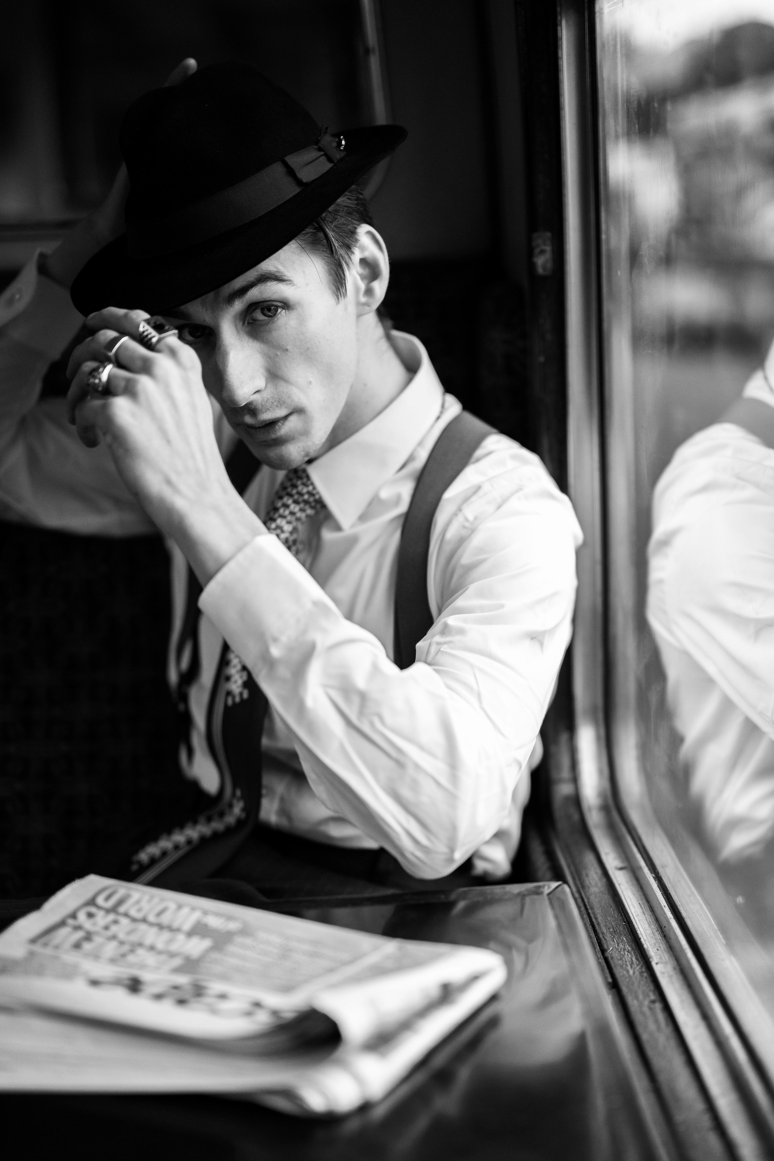 Model Lee Jay Hoy is photographed on The Creativity Hub's Murder on the Express photoshoot inside a train carriage next to a window, adjusting his hat and looking directly towards the camera. He's wearing smart vintage clothing and there is also a ne