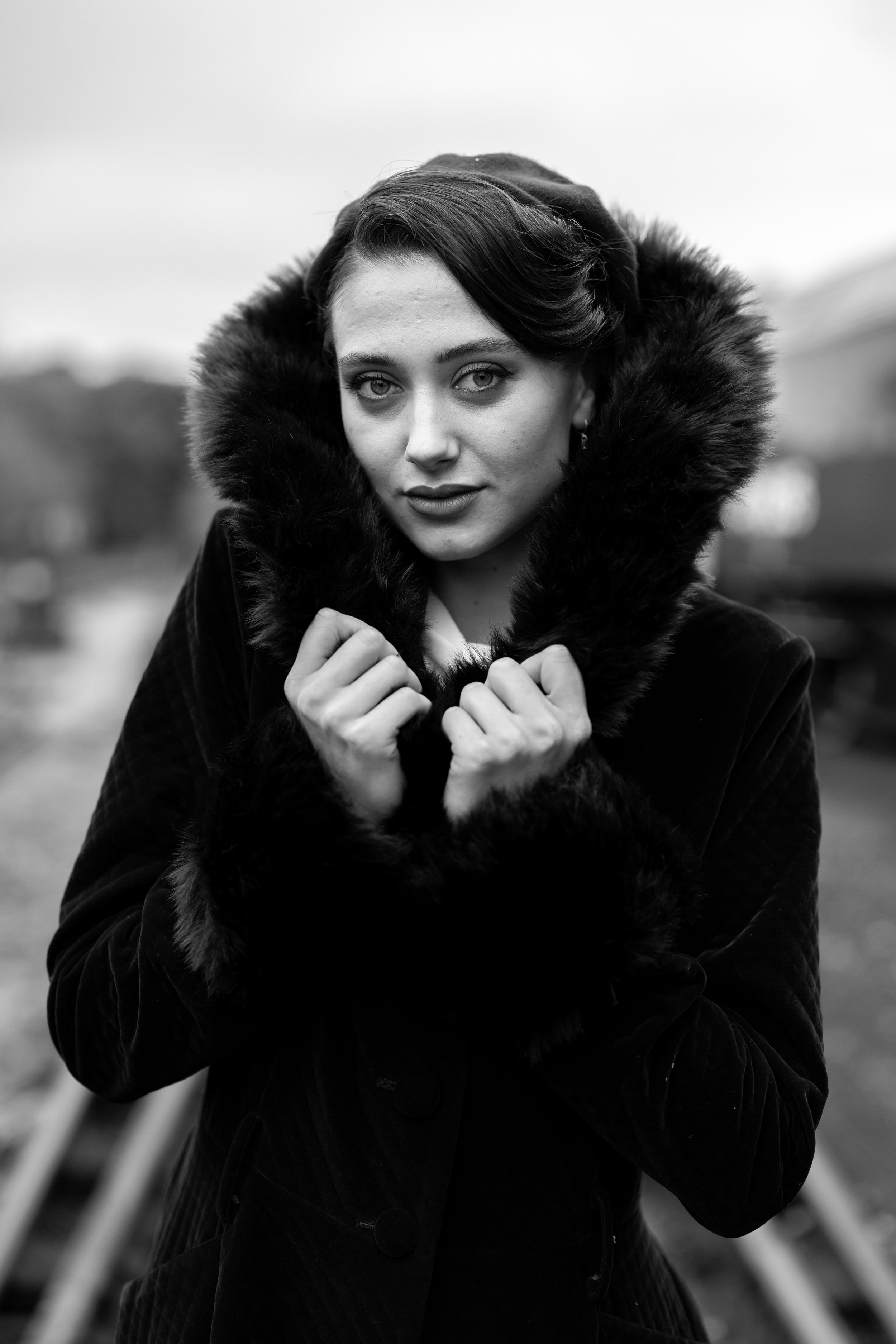 Model Molly Shaw-Downie is photographed on The Creativity Hub's Murder on the Express photoshoot stood on train tracks and pulling up the fur of a vintage coat collar around her face. The picture has been converted to B&W.
