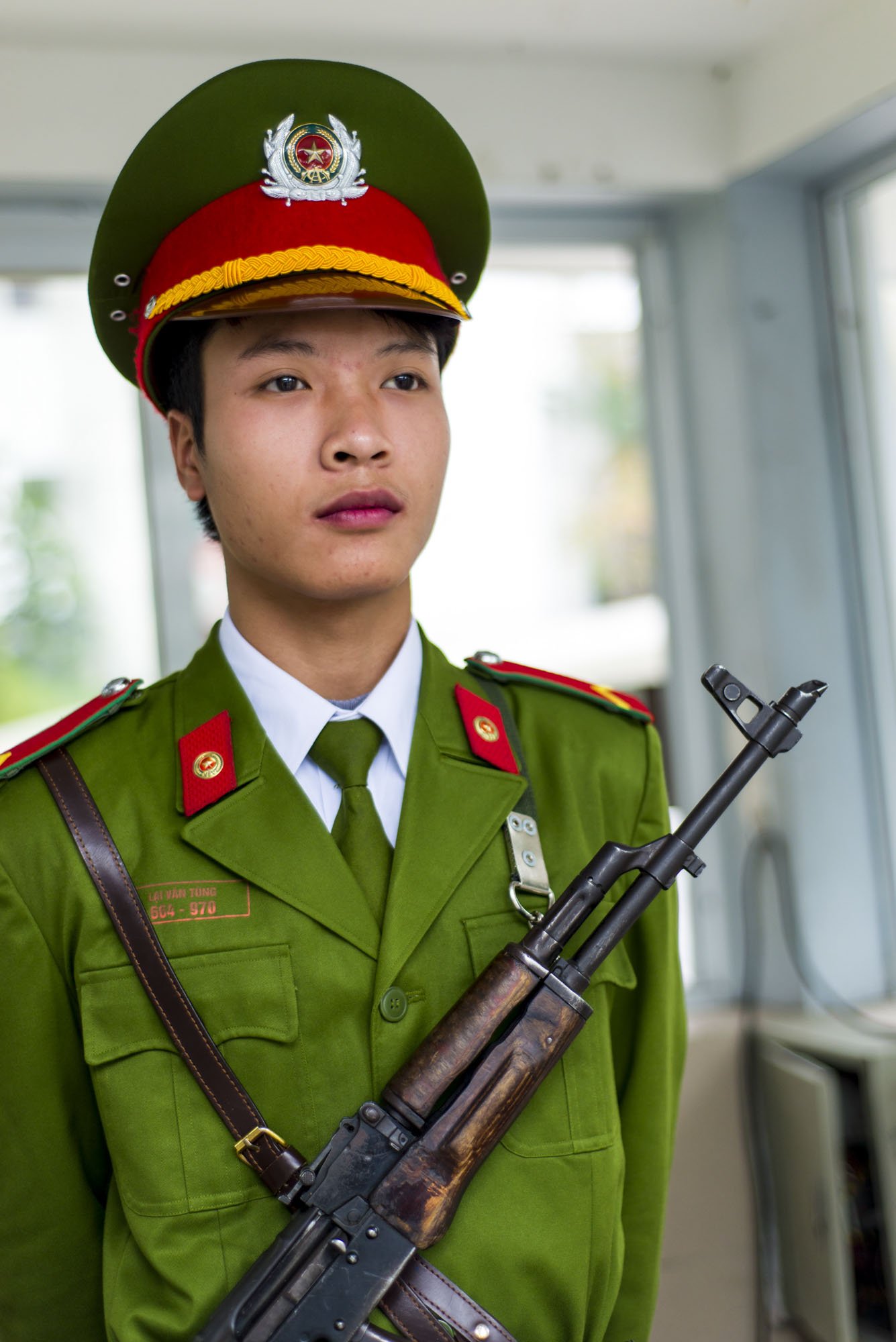 This is a photo of a soldier stood in a guard station in Hanoi, Vietnam in green military uniform wearing a rifle across his chest.