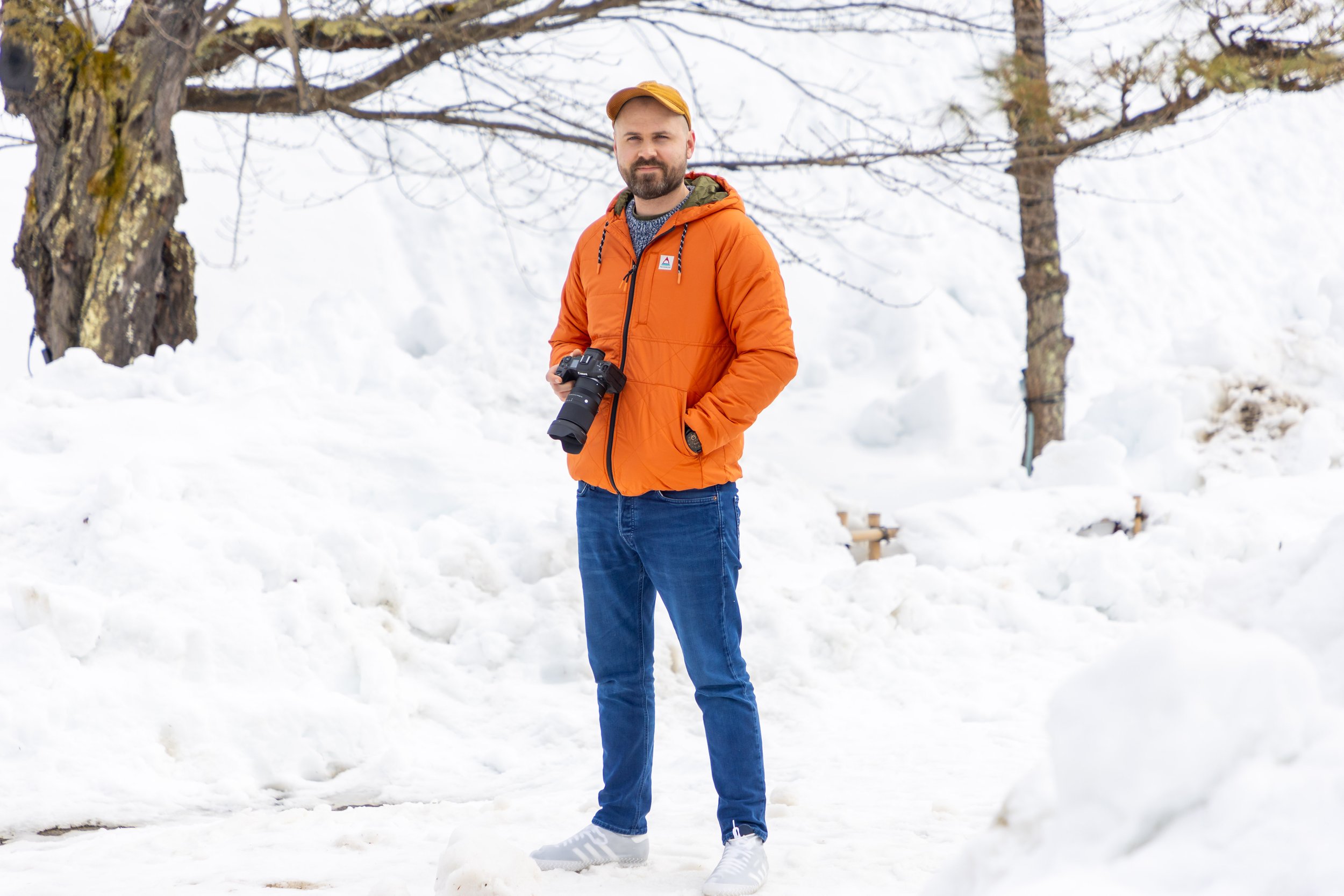 Professional photographer Dan Mold is pictured wearing a bright orange coat holding a Canon camera surrounded by snow, taken in Aizu Japan.