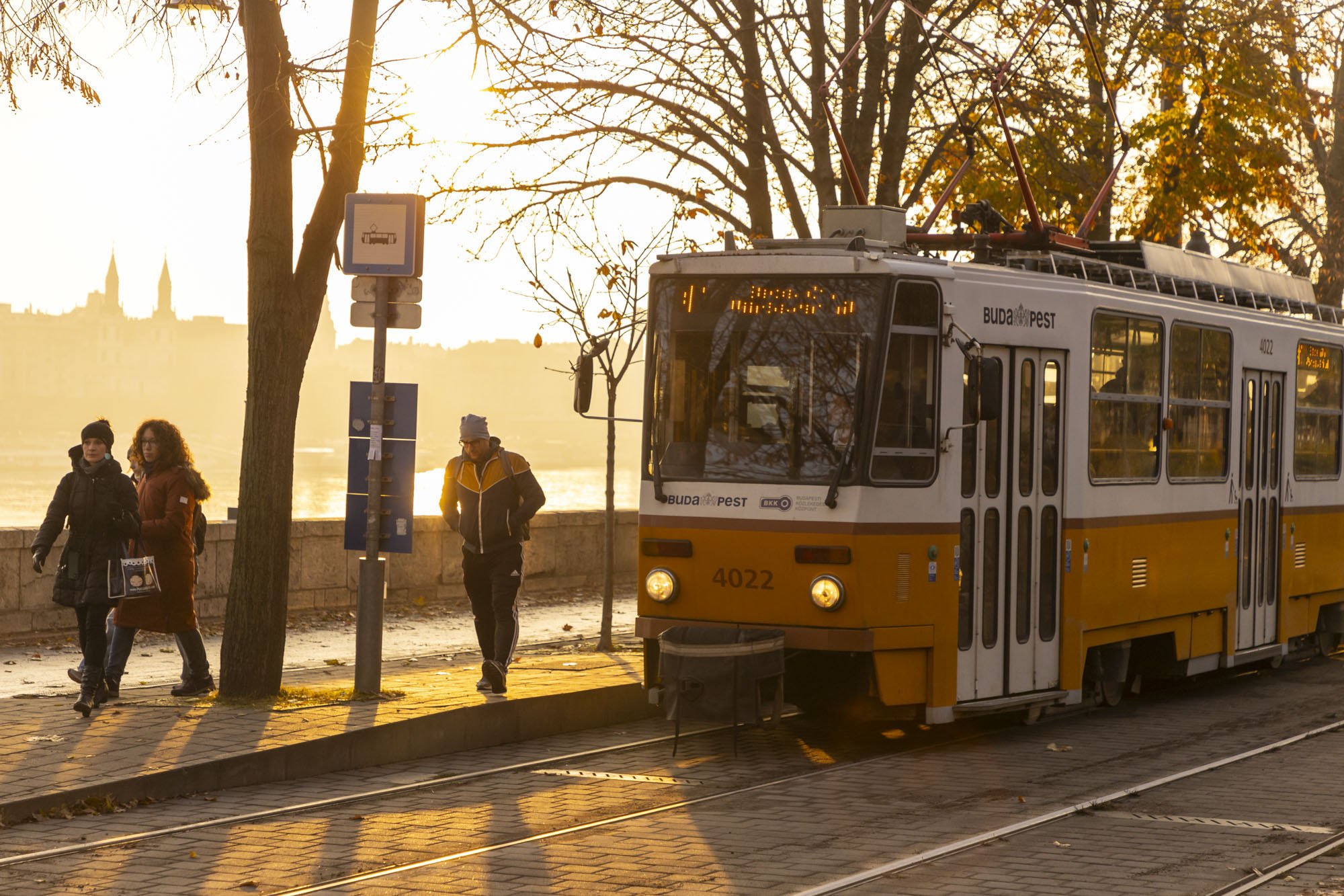 A city scene of Budapest with a tram and pedestrians walking past at sunrise with warm golden tones coming from the sun behind.