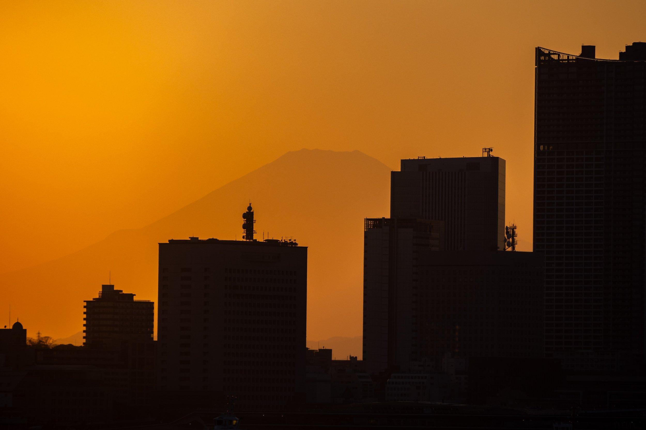 This image was taken with a very long focal length and zoomed in to compress the perspective so that the skyline of Yokohama's skyscrapers are silhouetted against the sunset but also the iconic shape of Mt Fuji is faintly seen behind the buildings.