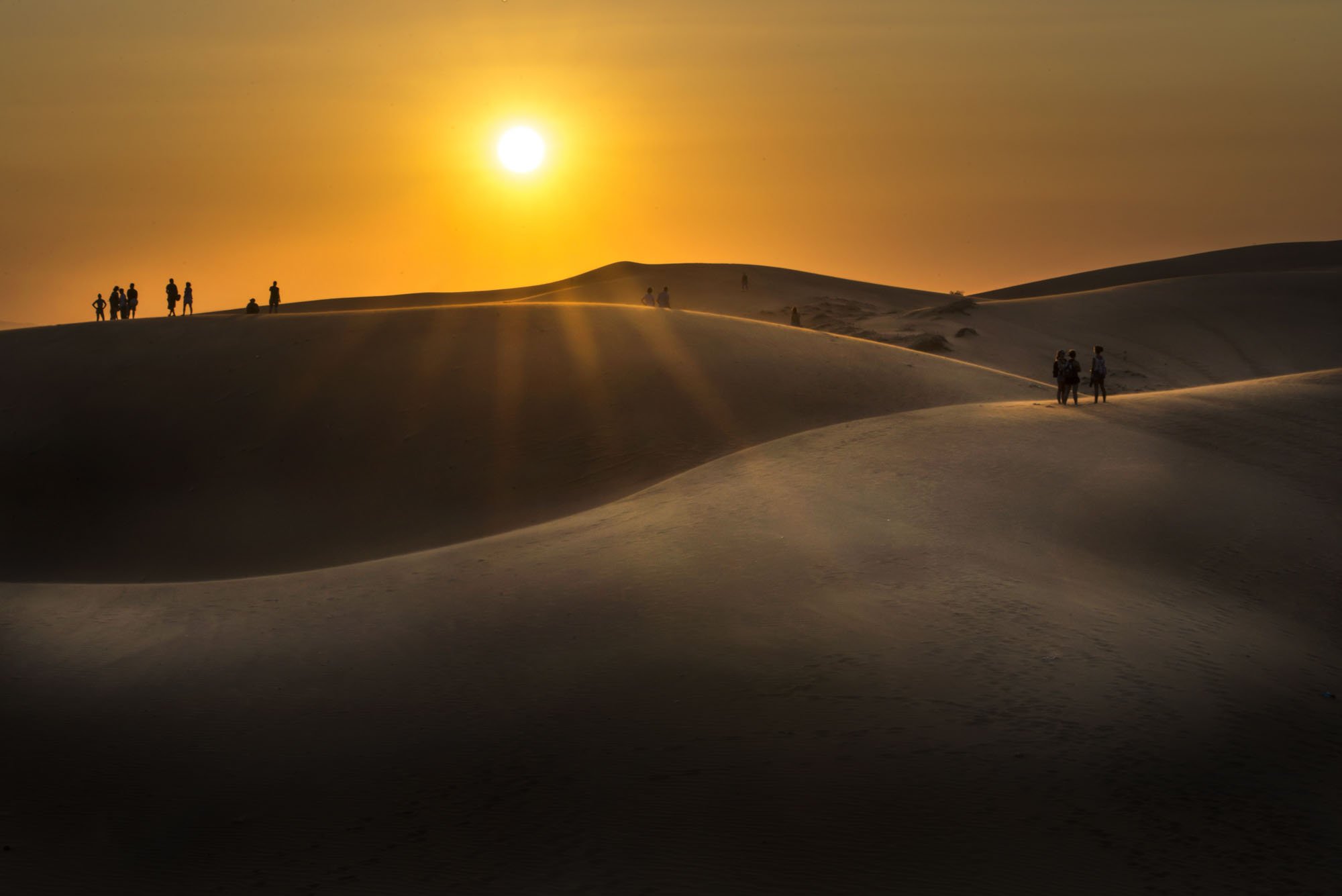 Sand dunes of Mui Ne in Vietnam at sunset with the low sun picking out the ridges of the dunes beautifully. A few people are on the dunes and have been turned into silhouettes.