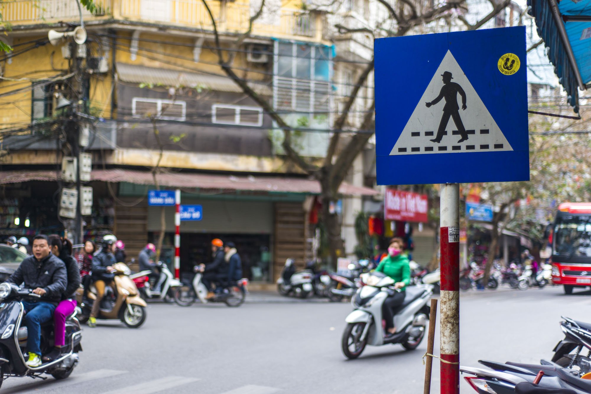 A street scene of a busy intersection in Hanoi, Vietnam with scooter riders whizzing past in the distance. In the foreground there is a blue pedestrian crossing sign where the pictured man crossing is wearing a hat.