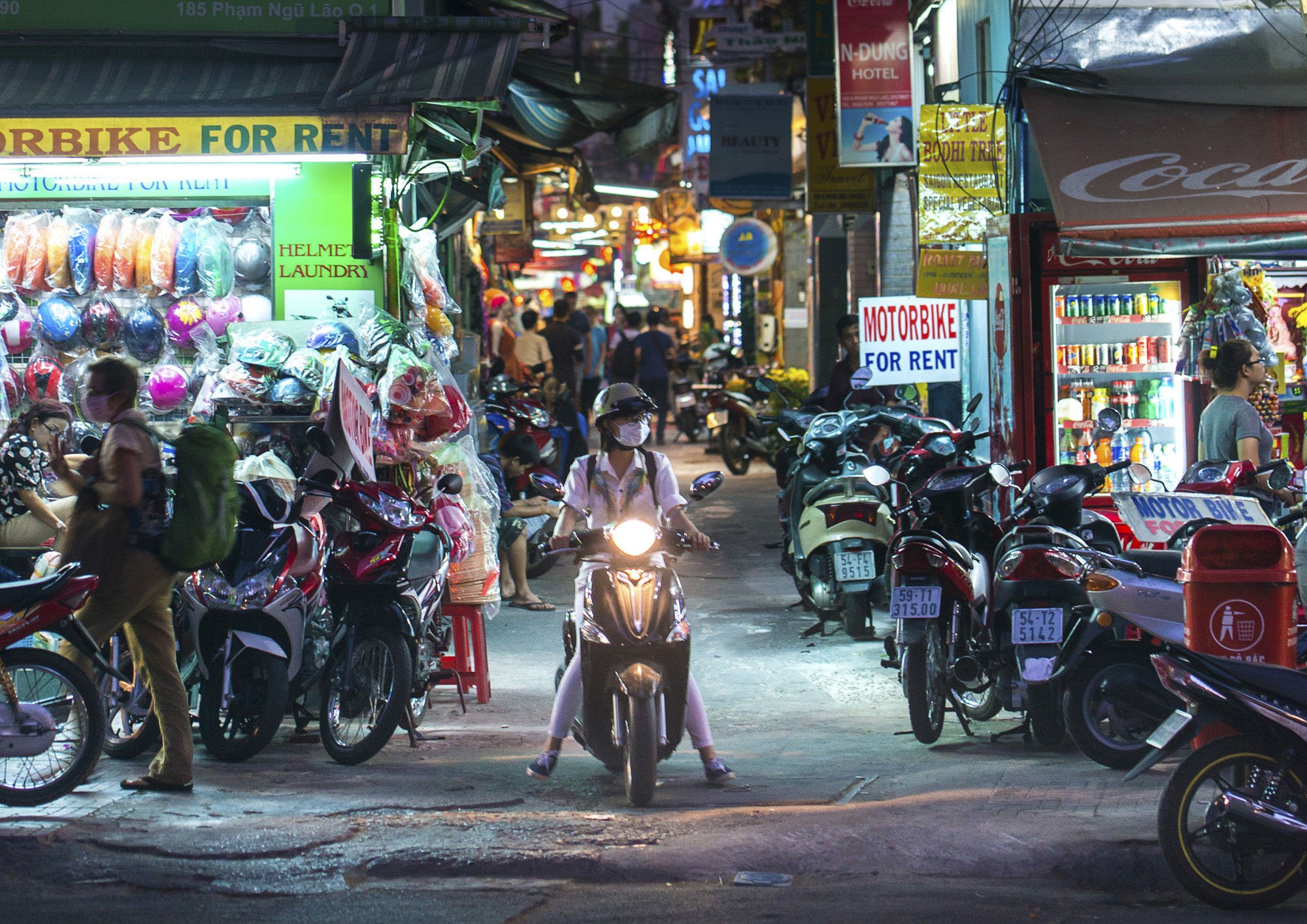 A night scene of Ho Chi Minh city with a woman on a scooter motorbike riding down a small crowded backstreet.