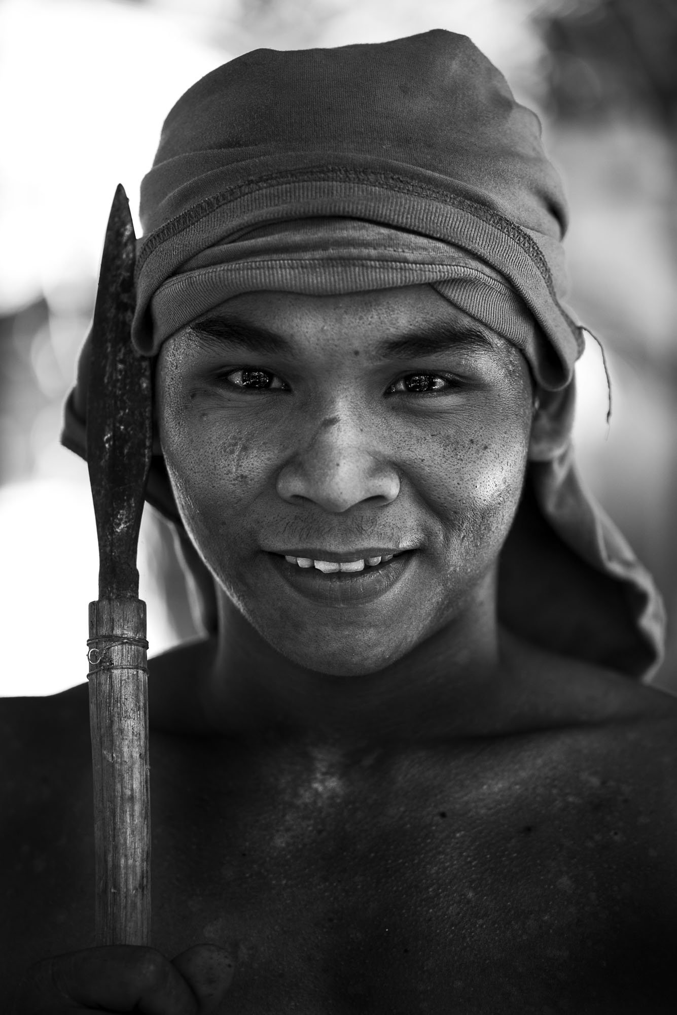 This is a portrait of a man taken in Philippines in a tribal setting with a shirt on his head and holding a spear close to his face. The image has been converted to black and white.
