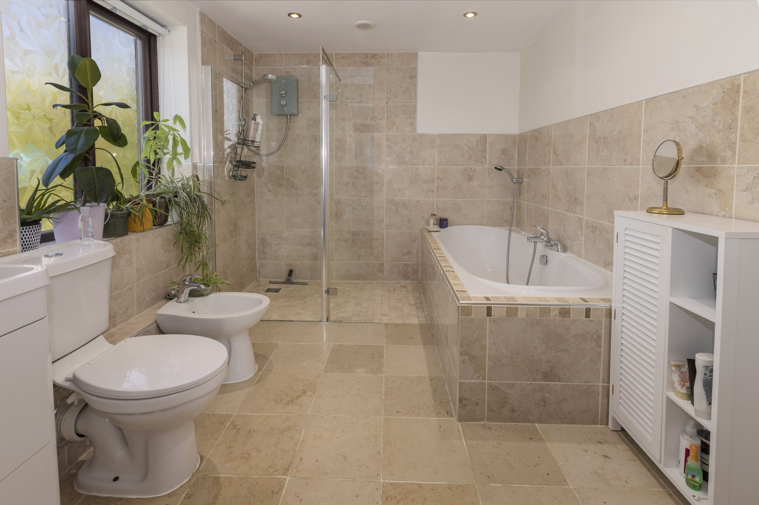 A bathroom with cream tiles which is light and bright with green plants on the window shelf and a bath in the corner.