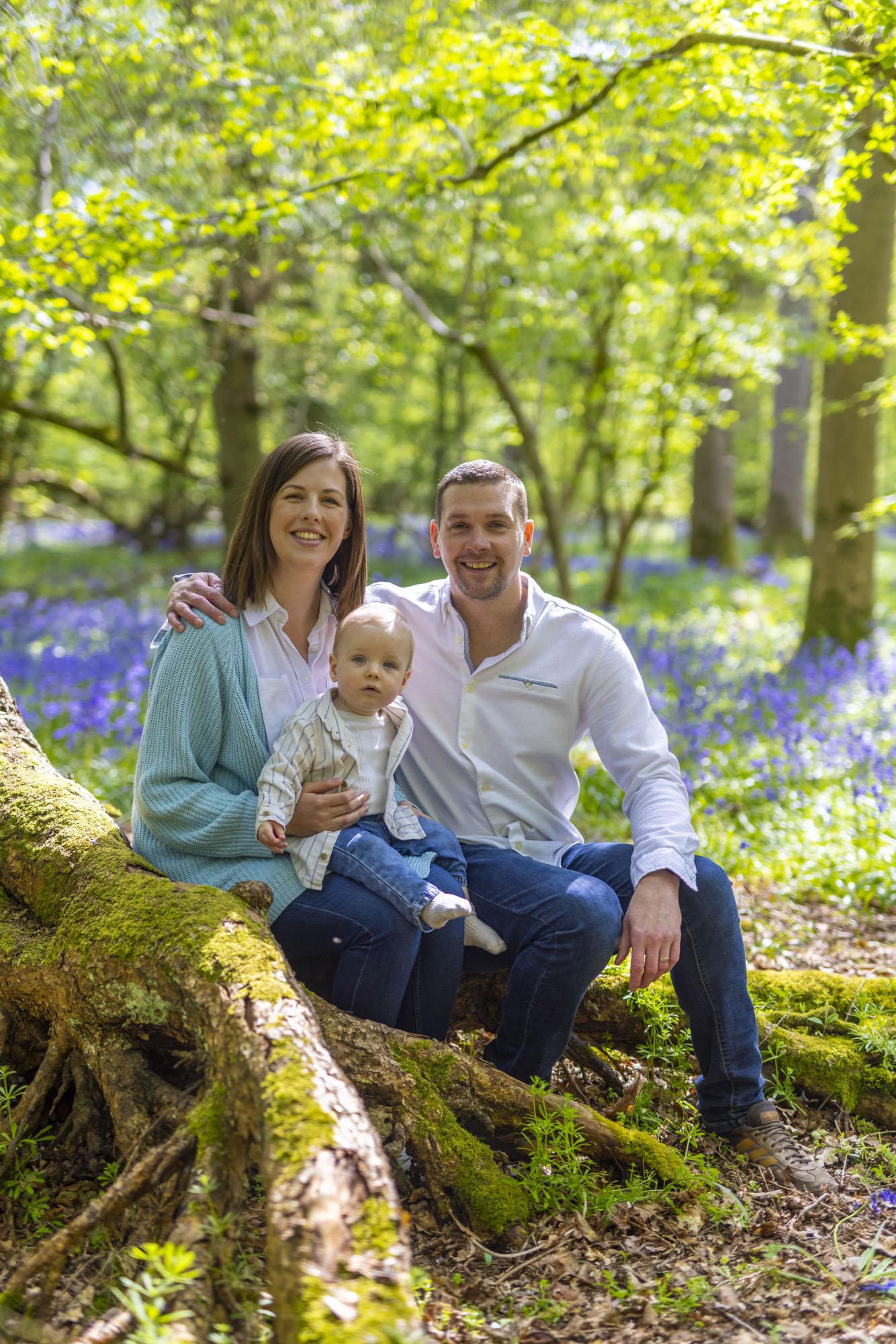 A family group shot of a mother and father with their young baby boy on the mothers lap. They are in a forest perched on a tree trunk with bluebells carpeting the forest floor. It's a bright sunny day but they are in the shade of the trees and smilin