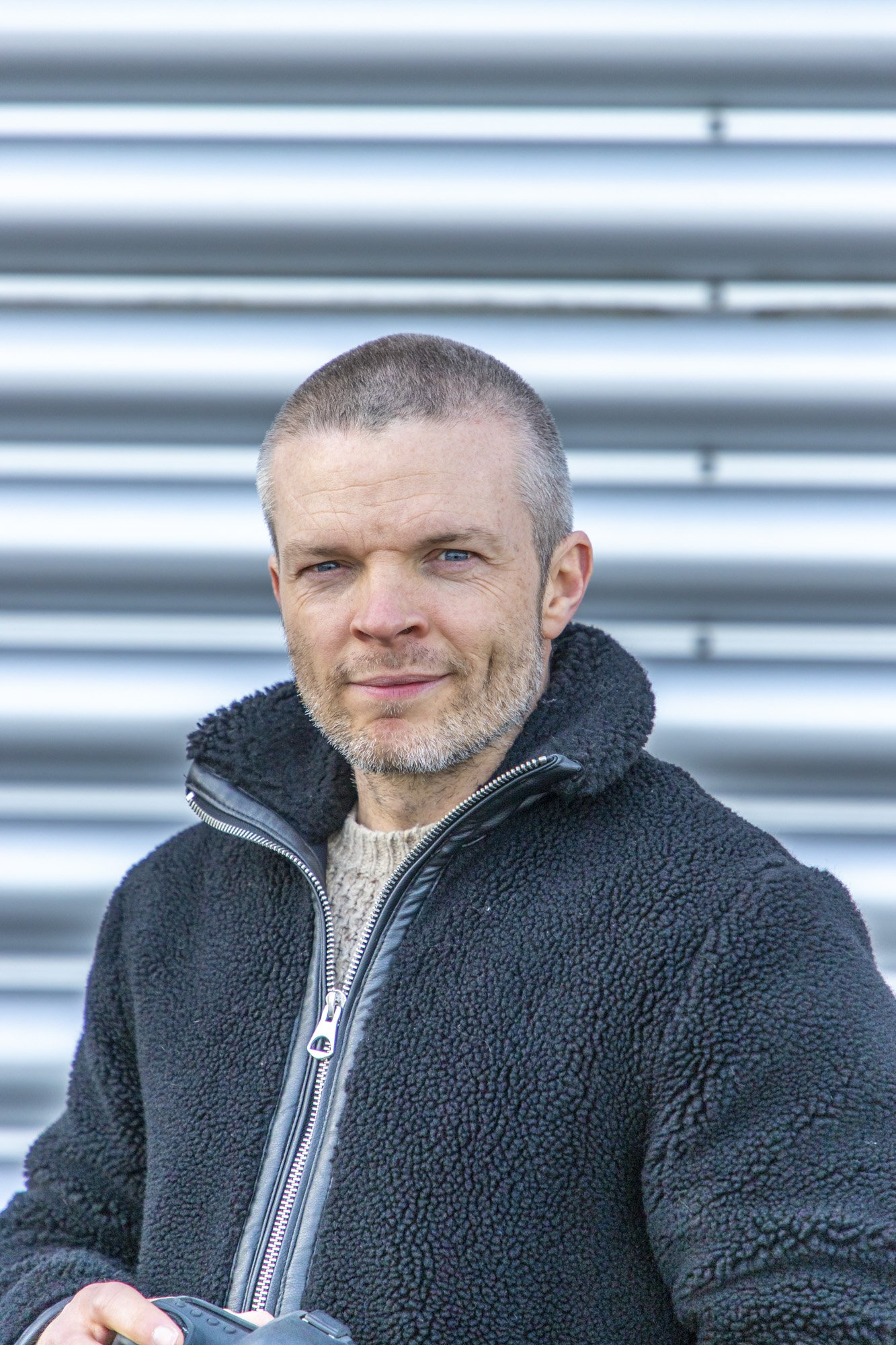 Professional automotive car photographer Alan Kenny poses for a headshot in front of metal garage doors holding a camera in his hands.