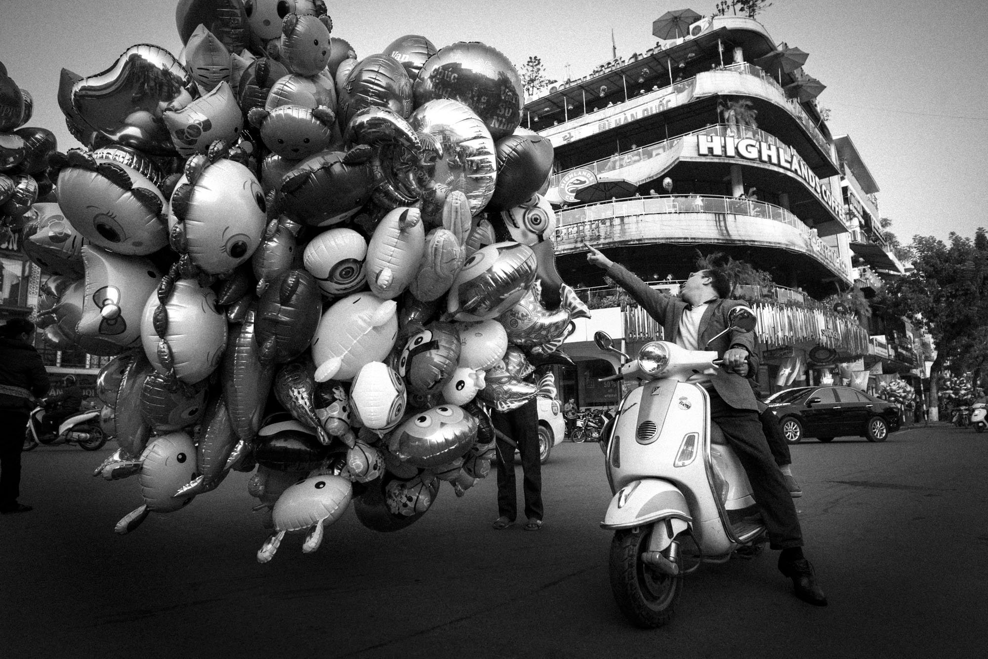 A man stops on a busy street in Hanoi to buy balloons from a balloon salesman. It's a comical shot with him pictured on a scooter pointing towards a huge mass of balloons which takes up half the frame.
