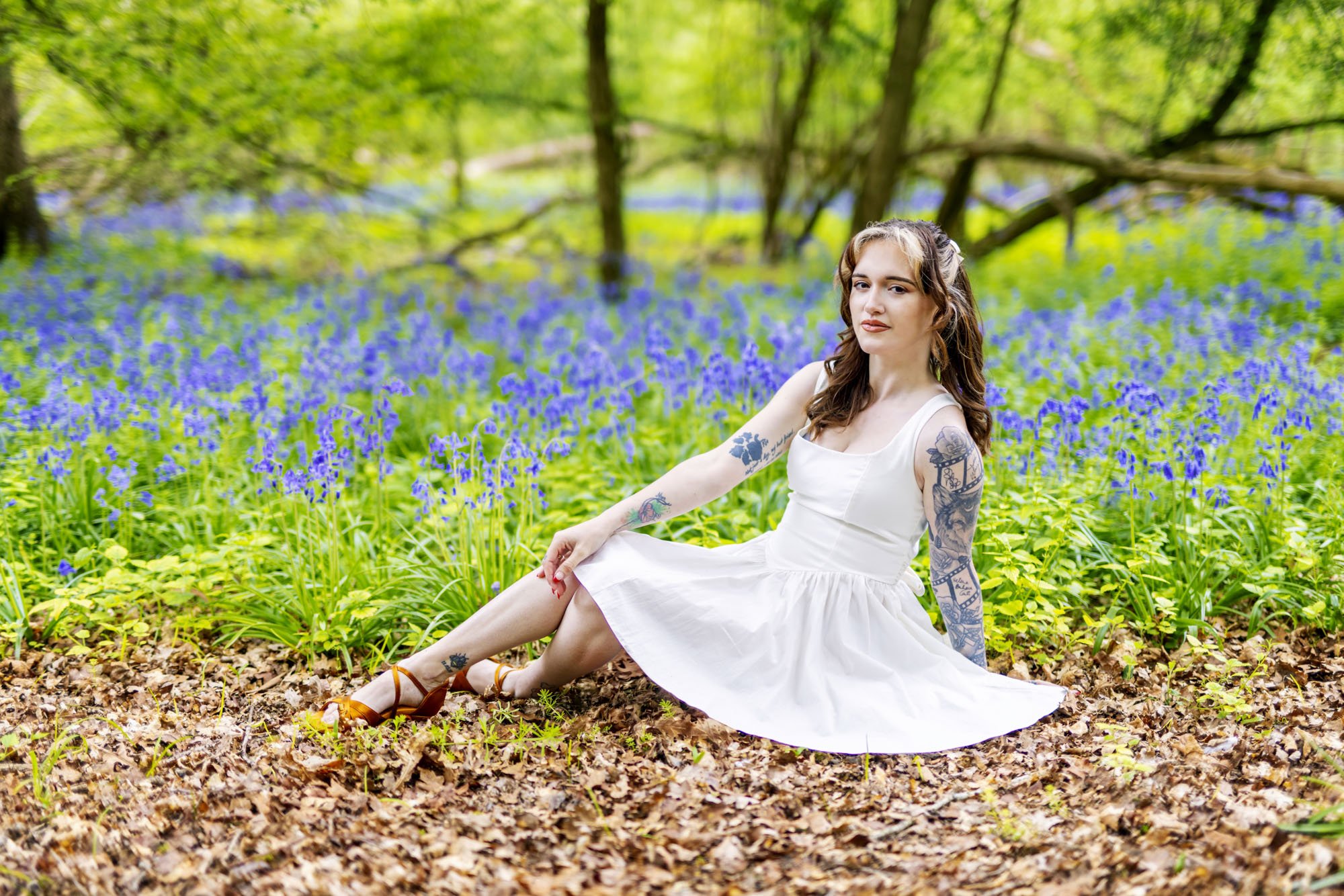 Model Megan Annie is pictured sat on a forest floor of a bluebell woods wearing a knee-length white dress.