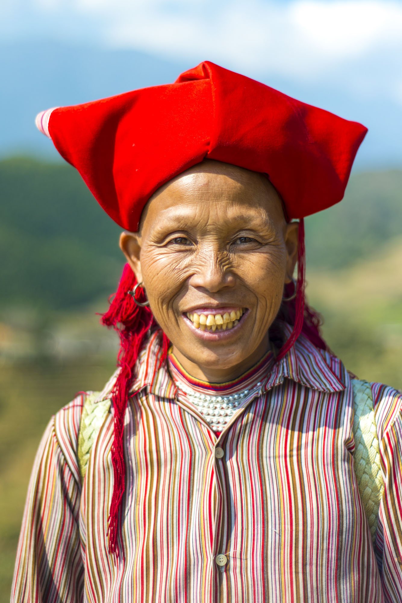 A woman is photographed in Sa Pa, Vietnam wearing a striped red shirt and a red headpiece. She is smiling and looking directly towards the camera. The portrait was taken outdoors with the rice fields in the background blurred with a shallow depth of