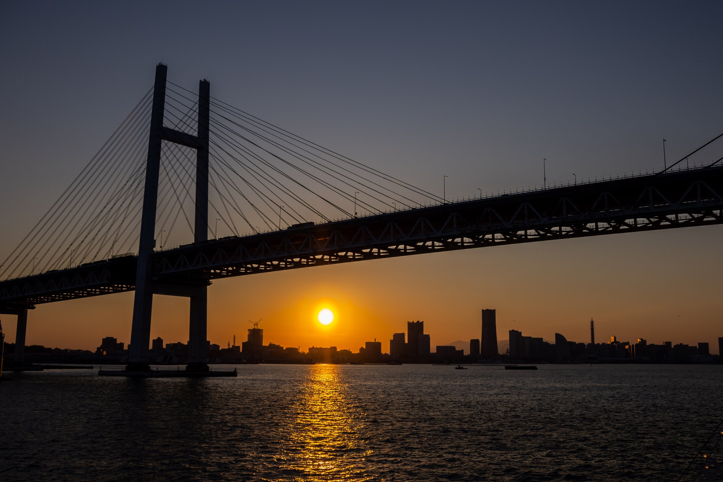 A sunset scene taken in Yokohama, Japan with the sun setting over the city. This image was taken from a ferry and has just passed under a bridge which takes up most of the frame - the city skyline and sun are pictured underneath the bridge with the i