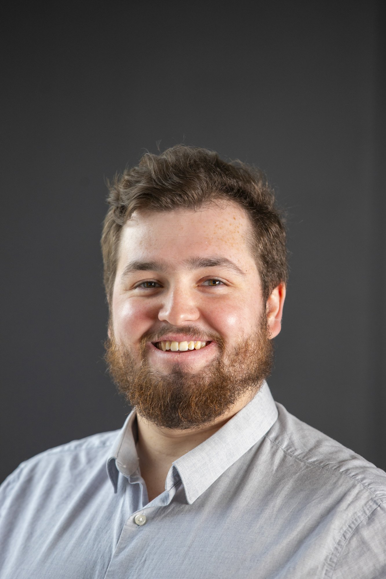 A man wearing a white shirt with dark hair and a beard poses smiling towards the camera for a headshot in a studio with a black background.