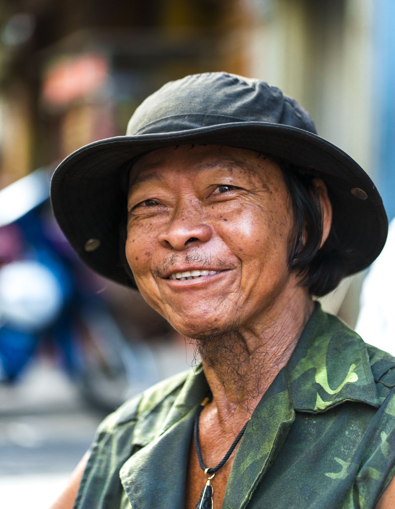 A Vietnamese man is photographed on the streets. He is wearing casual military green clothing and is looking towards the camera and smiling.