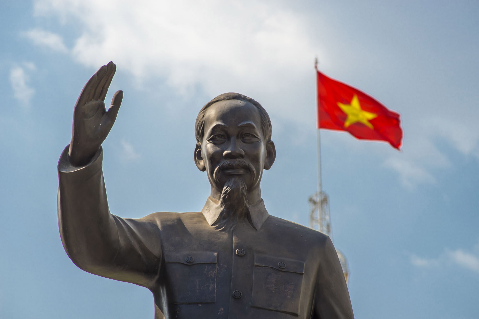A statue in Ho Chi Minh city, Vietnam of Ho Chi Minh saluting with the Vietnamese flag being flown behind. It's a sunny day with blue sky and a cloud seen behind.