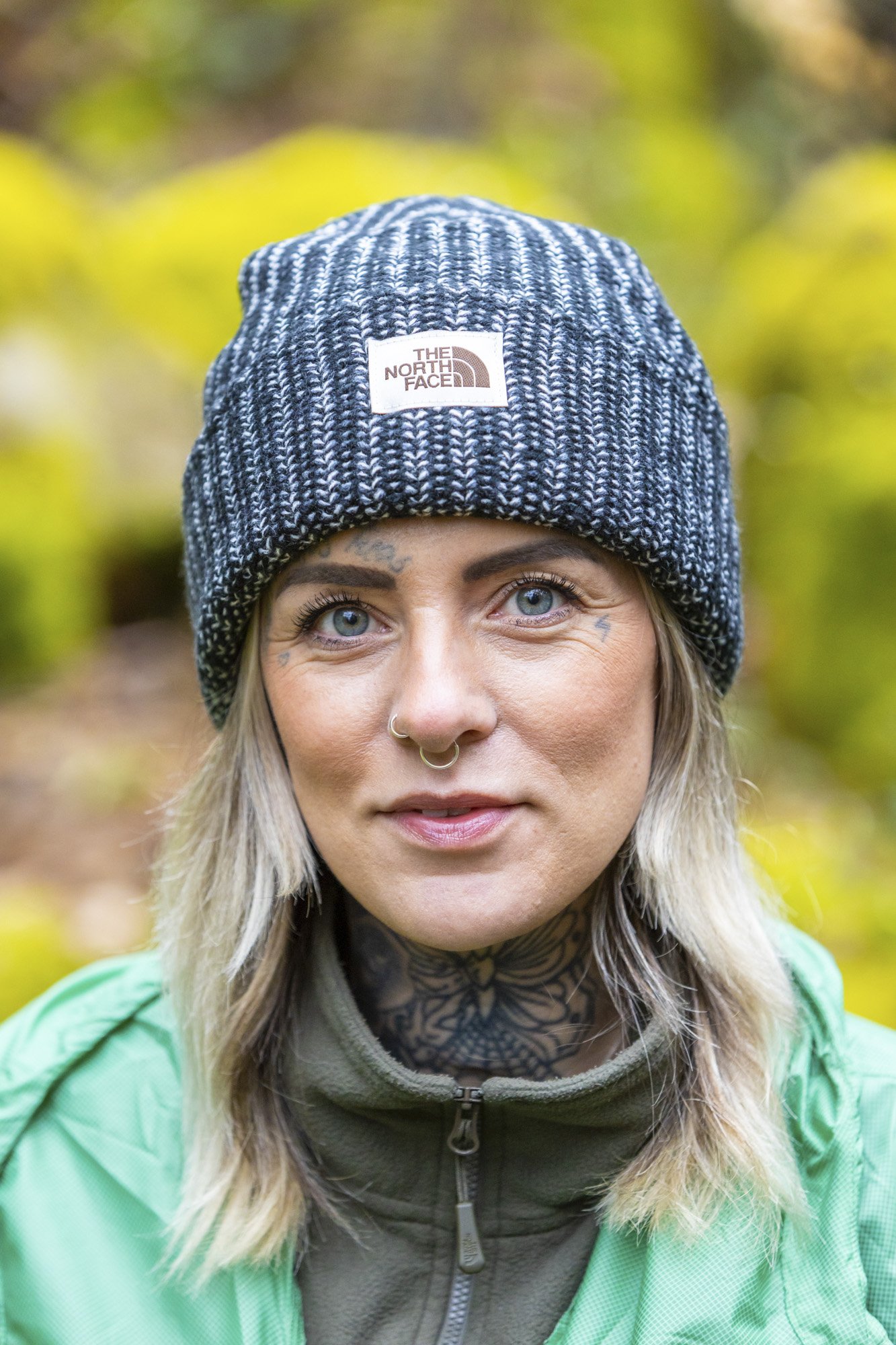An outdoorsy woman poses for a headshot outside in a nature park wearing a green waterproof jacket and grey wool beanie hat. Behind her the mossy background has been blurred out.