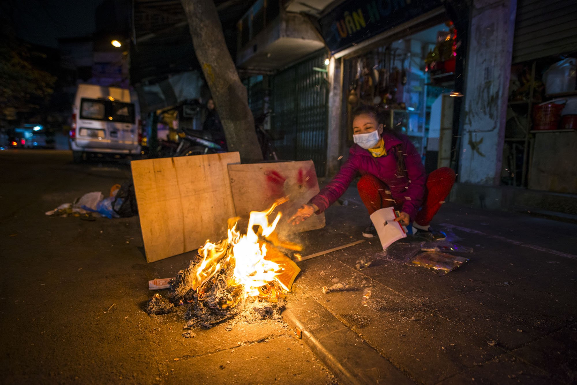 A woman pictured on the streets of Hanoi, Vietnam at night throwing fake money into a fire on the street. She's wearing red and a face mask.