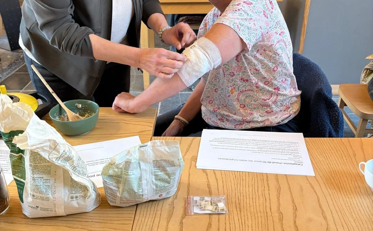 Une personne se fait soigner le bras par une herboriste, dans un cadre intérieur. La personne assise porte un t-shirt à motifs floraux et a un pansement blanc sur le bras après qu'on lui a mis un cataplasme.