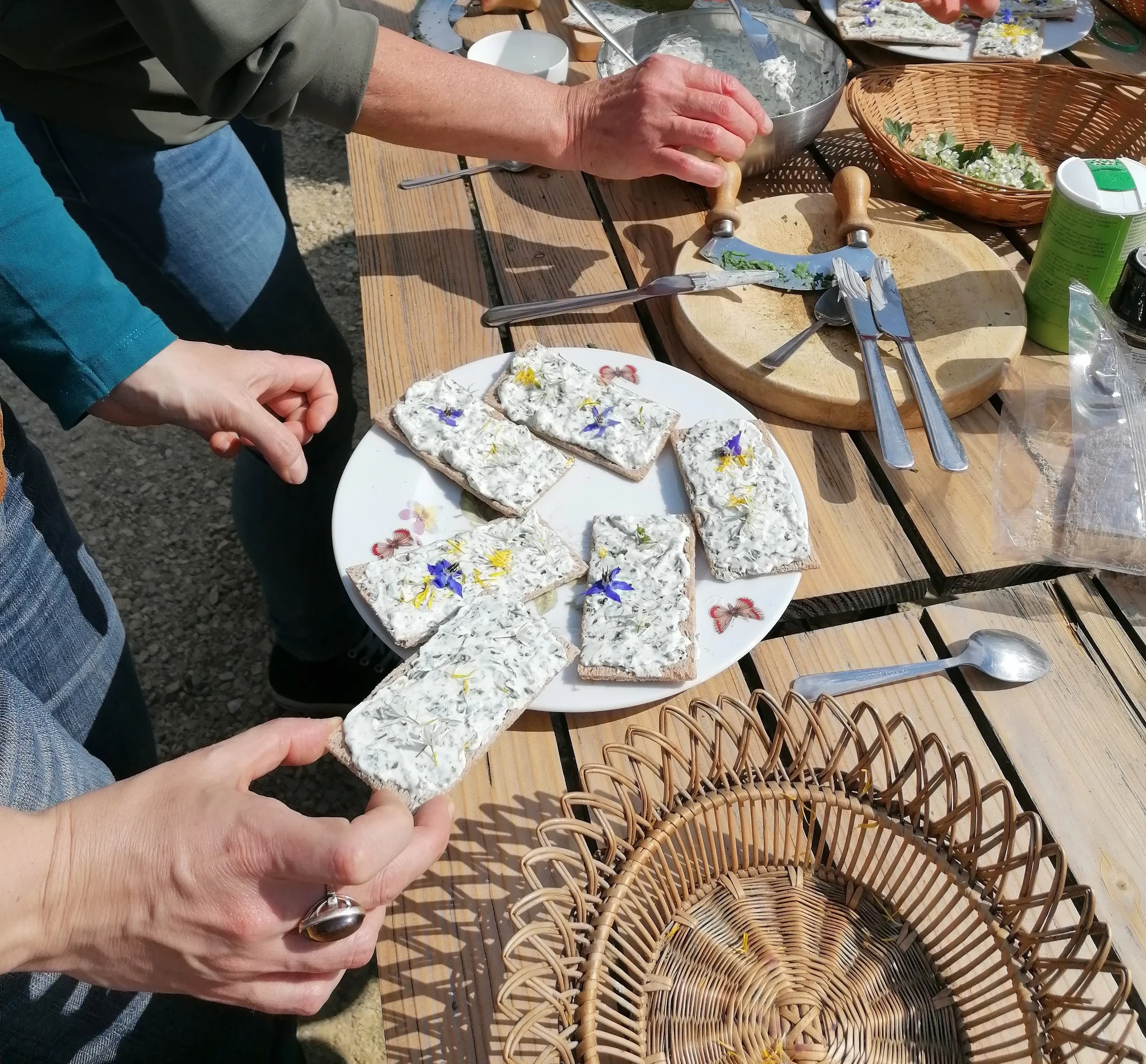 Mains tenant des biscuits garnis de fromage aux herbes avec des fleurs comestibles sur une table en bois, entourée d'ustensiles de cuisine et de nourriture.