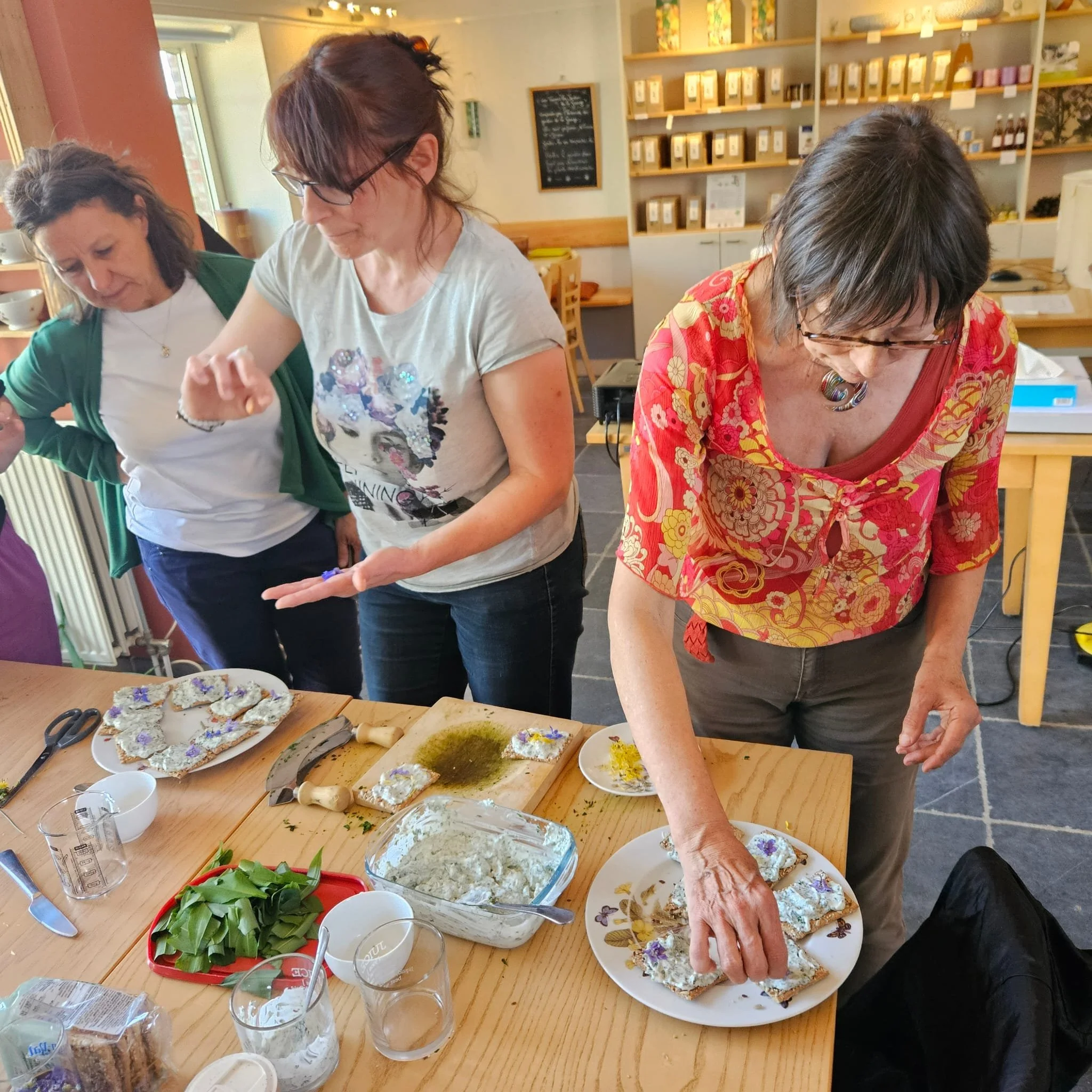 Trois femmes participent à un atelier de cuisine ou de préparation d'aliments, avec des plats et des ingrédients sur la table. Elles semblent décorer ou assembler des biscuits ou des amuse-bouches.
