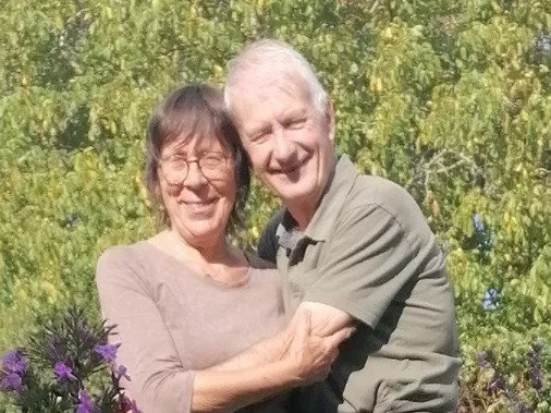Un couple d'âge mûr souriant, un homme et une femme, se tenant dans un jardin verdoyant avec des fleurs violettes.