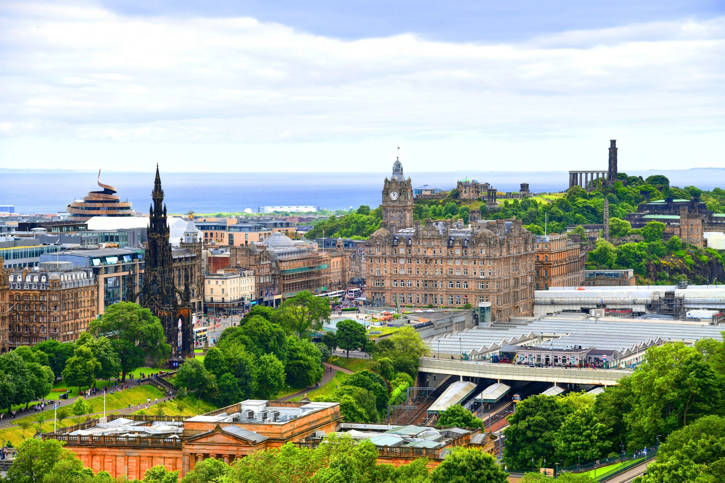 A panoramic view of Edinburgh, Scotland with historic buildings, a clock tower, lush green trees, and a hill with ruins in the background overlooking the sea.