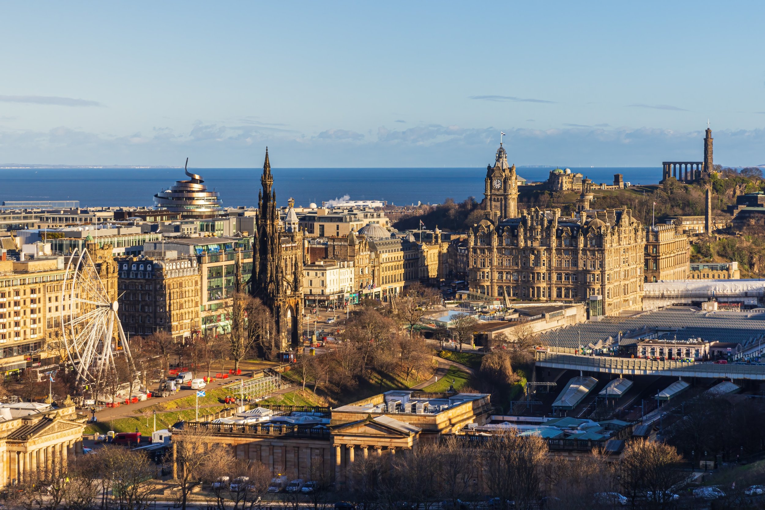 A panoramic view of Edinburgh, Scotland, featuring historic buildings, a Ferris wheel, and the sea in the background on a clear day.
