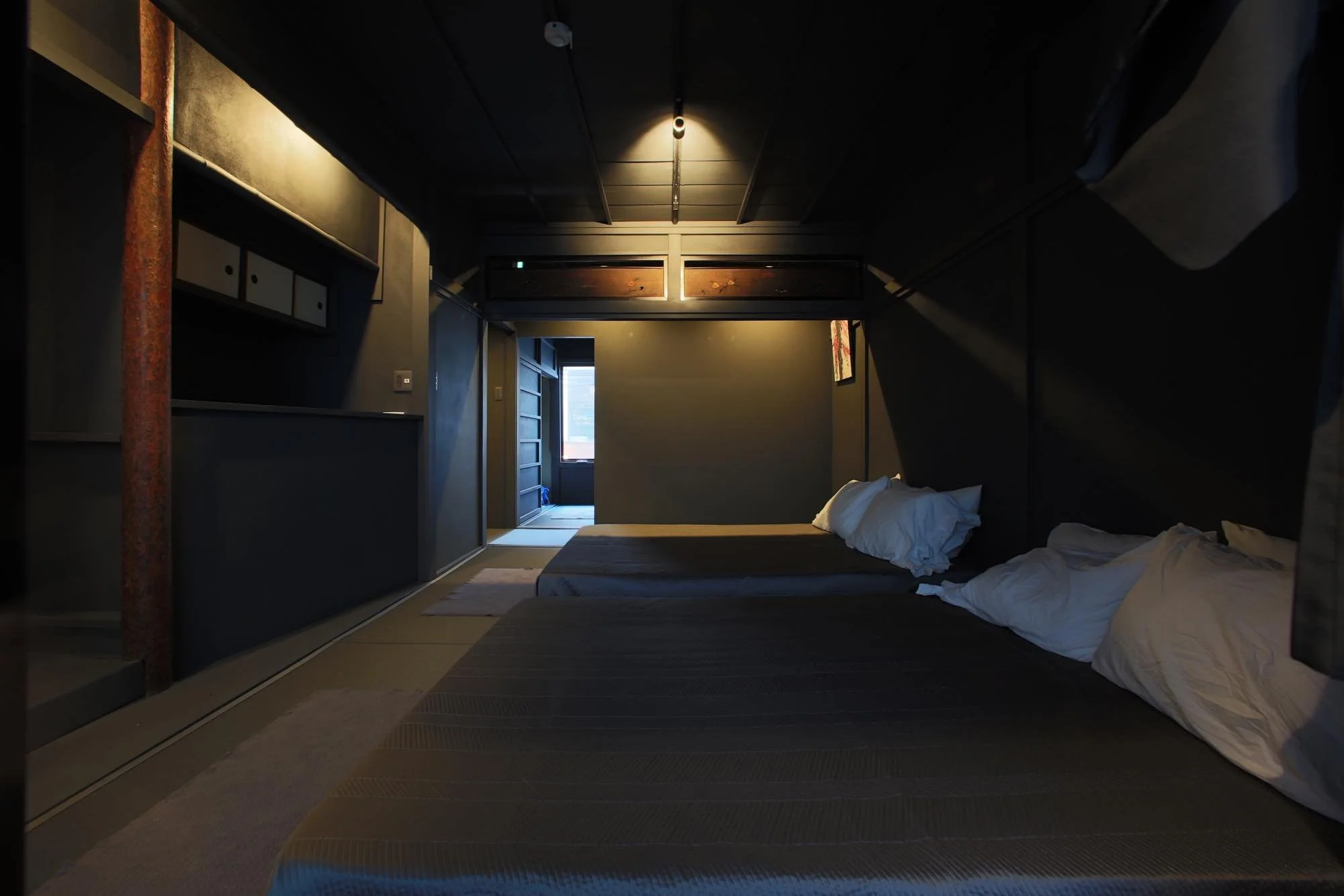 A cozy bedroom with two beds, one unmade with white sheets and pillows, in front of a dark wall with a window and a sliding door leading outside. There's a dark ceiling with a pendant light, a wardrobe, and a partial view of a painting.