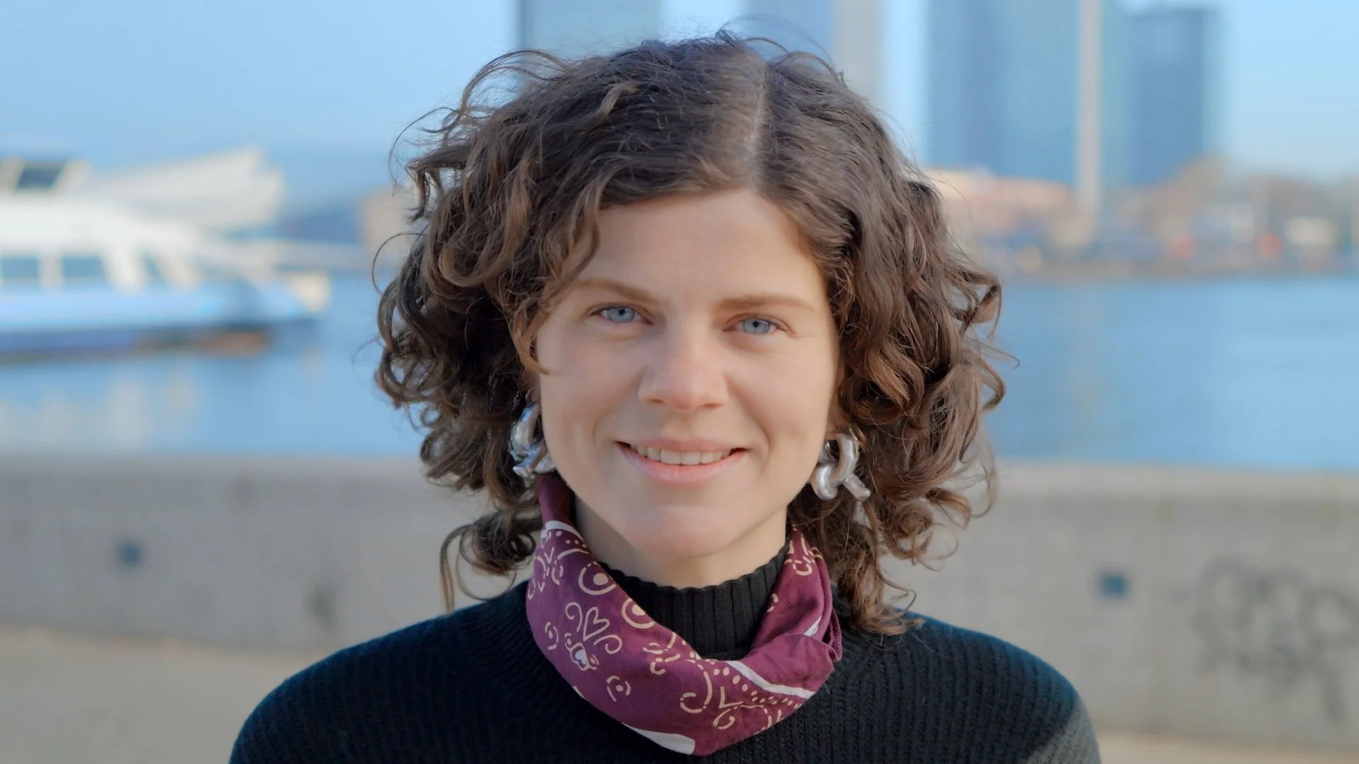 A woman with curly brown hair and blue eyes smiling outdoors near a body of water, with a city skyline in the background.