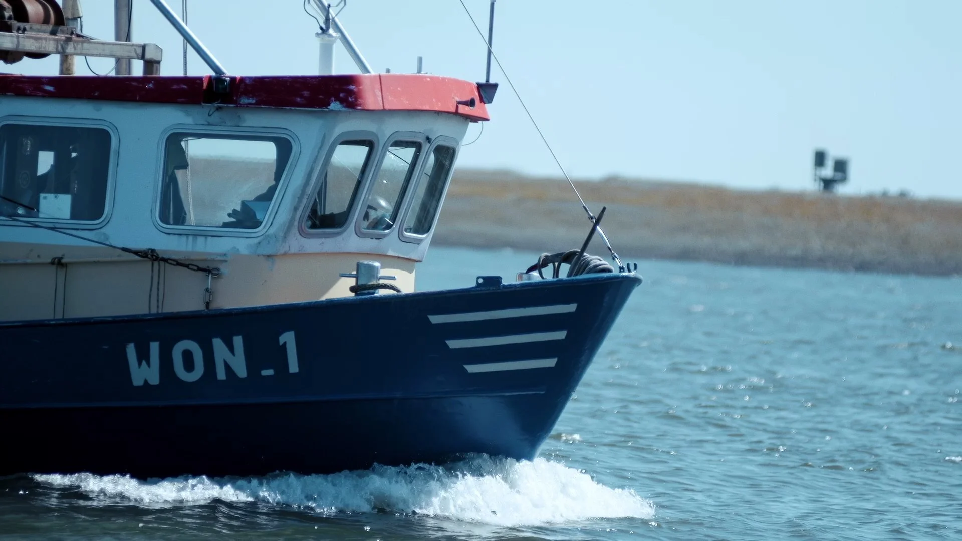 Close-up of a boat on water with the registration WON.1 visible on the hull, and land with some structures in the background.
