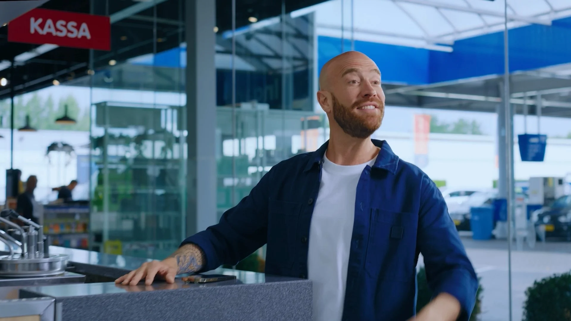 A man with a beard and tattoos on his arm is smiling at a cash register at a store entrance, with a parking lot visible outside.