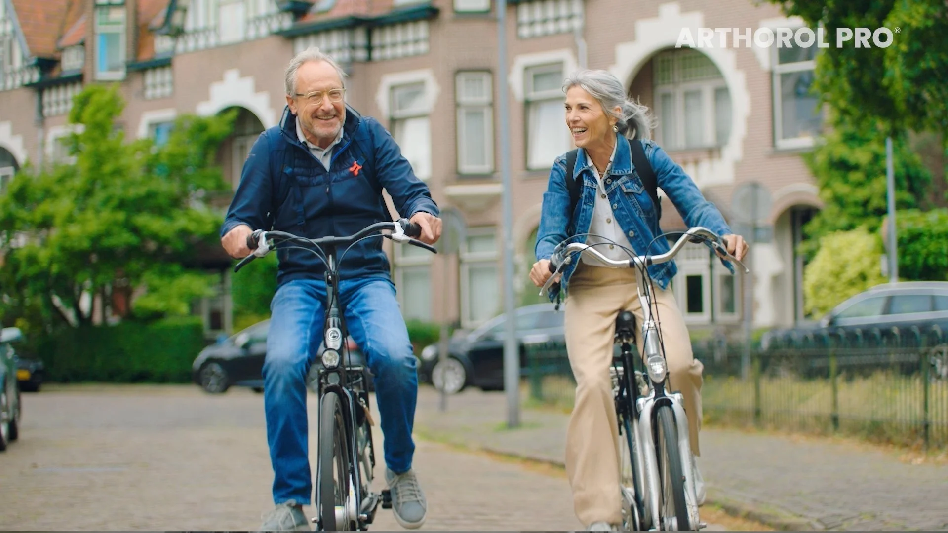 Older man and woman cycling on a city street, smiling and enjoying their ride with city buildings and greenery in the background.
