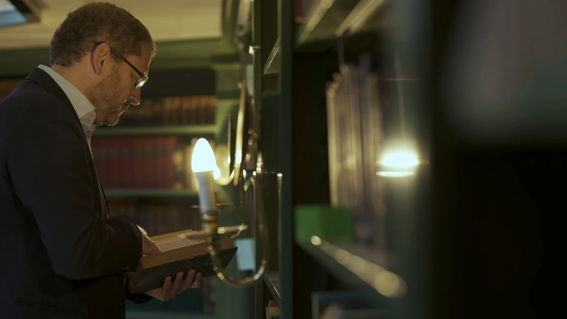 A man in a suit reading a book in a dimly lit library, illuminated by a table lamp.