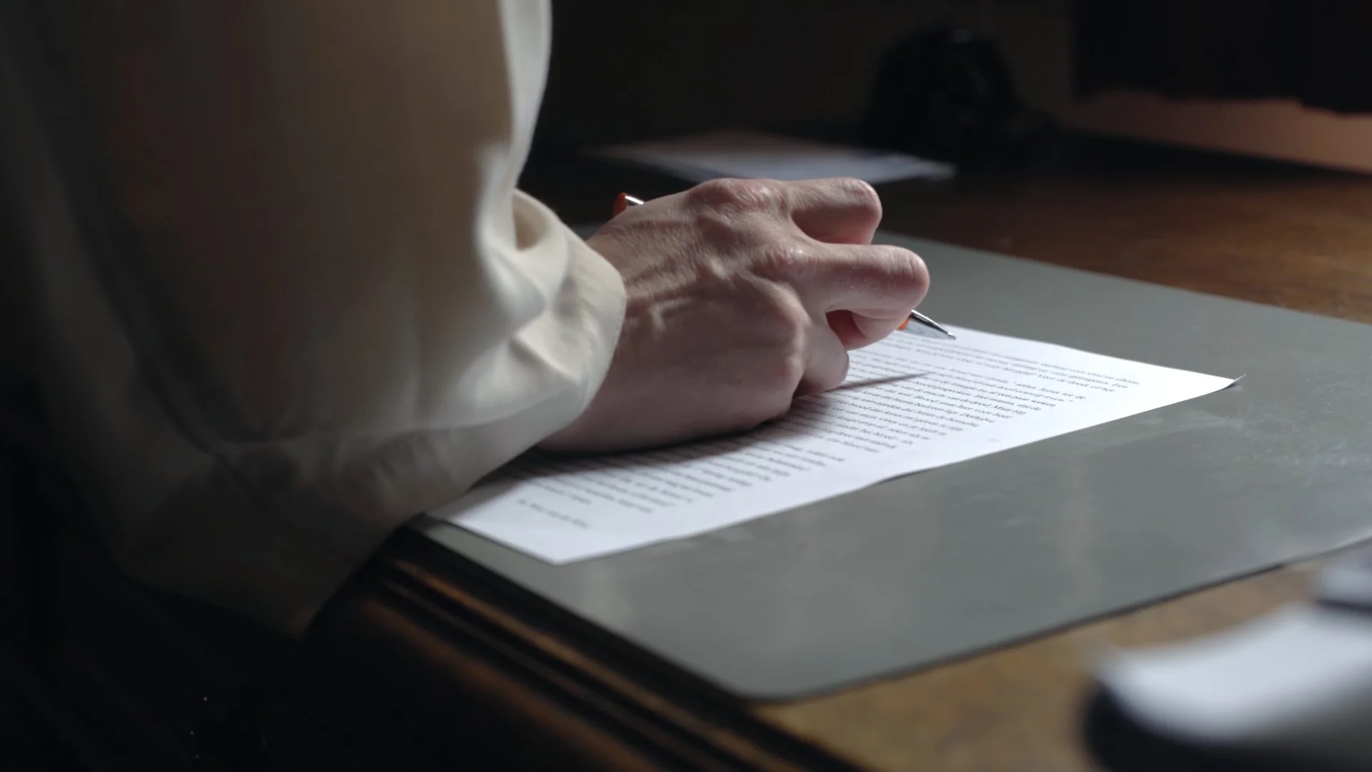 Person writing with a pen on a printed document, placed on a desk with a silver laptop and blurred objects in the background.