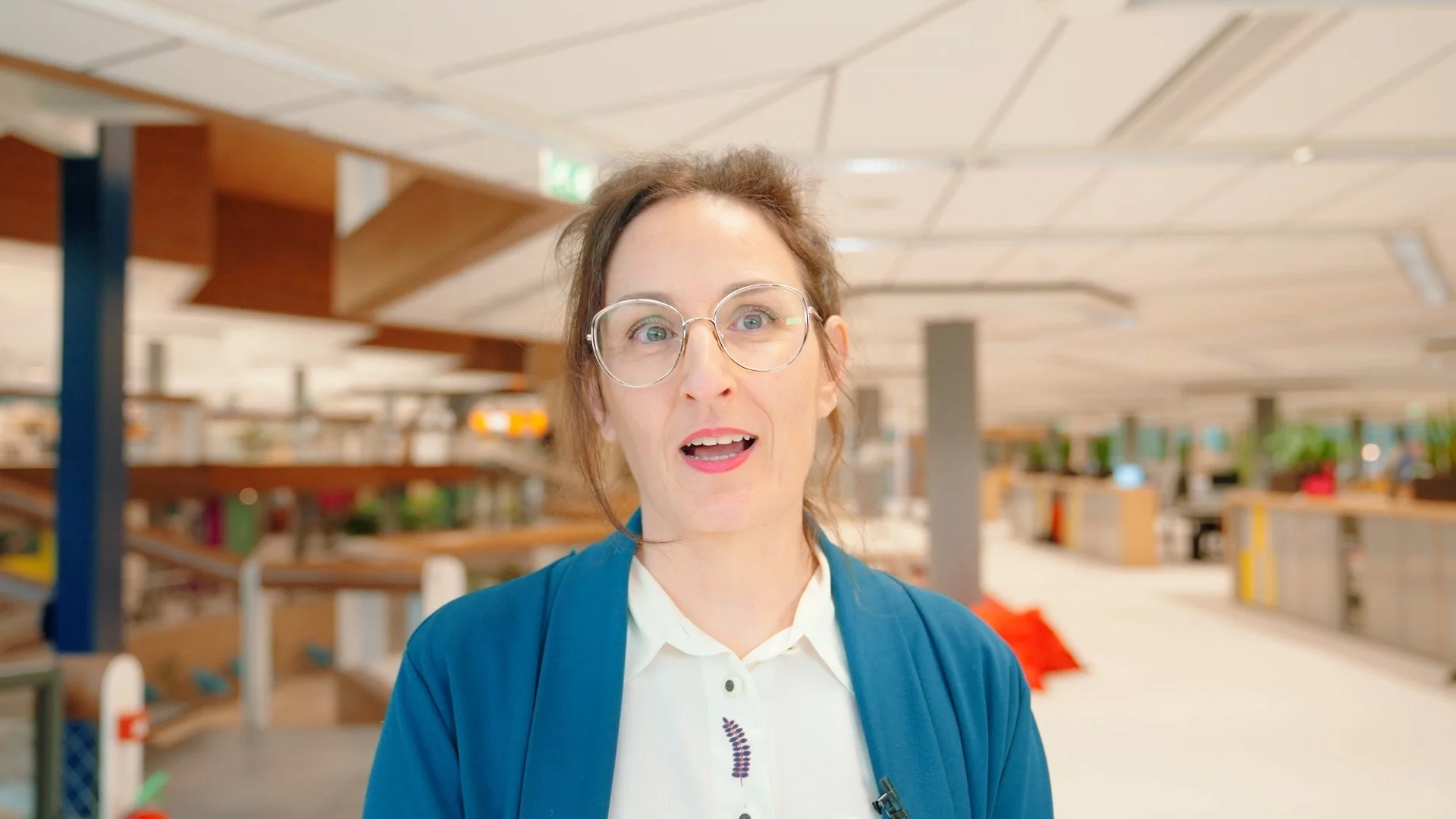 A woman with glasses, brown hair, and a blue jacket standing inside a brightly lit, modern indoor space with wooden panels and plants in the background.