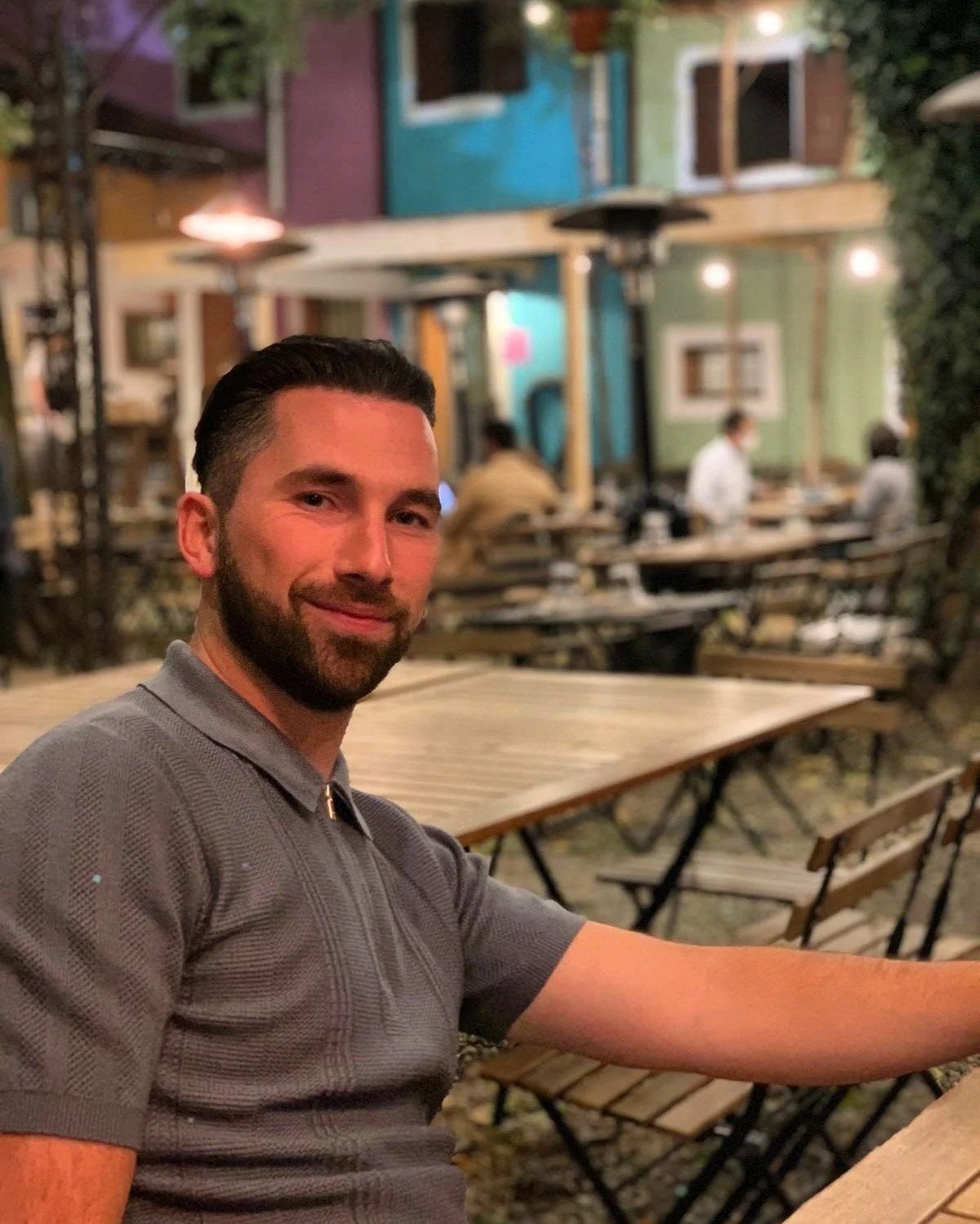 A smiling man with dark hair and a beard sitting at an outdoor patio with wooden tables and chairs, with people in the background and colorful buildings decorated with murals.
