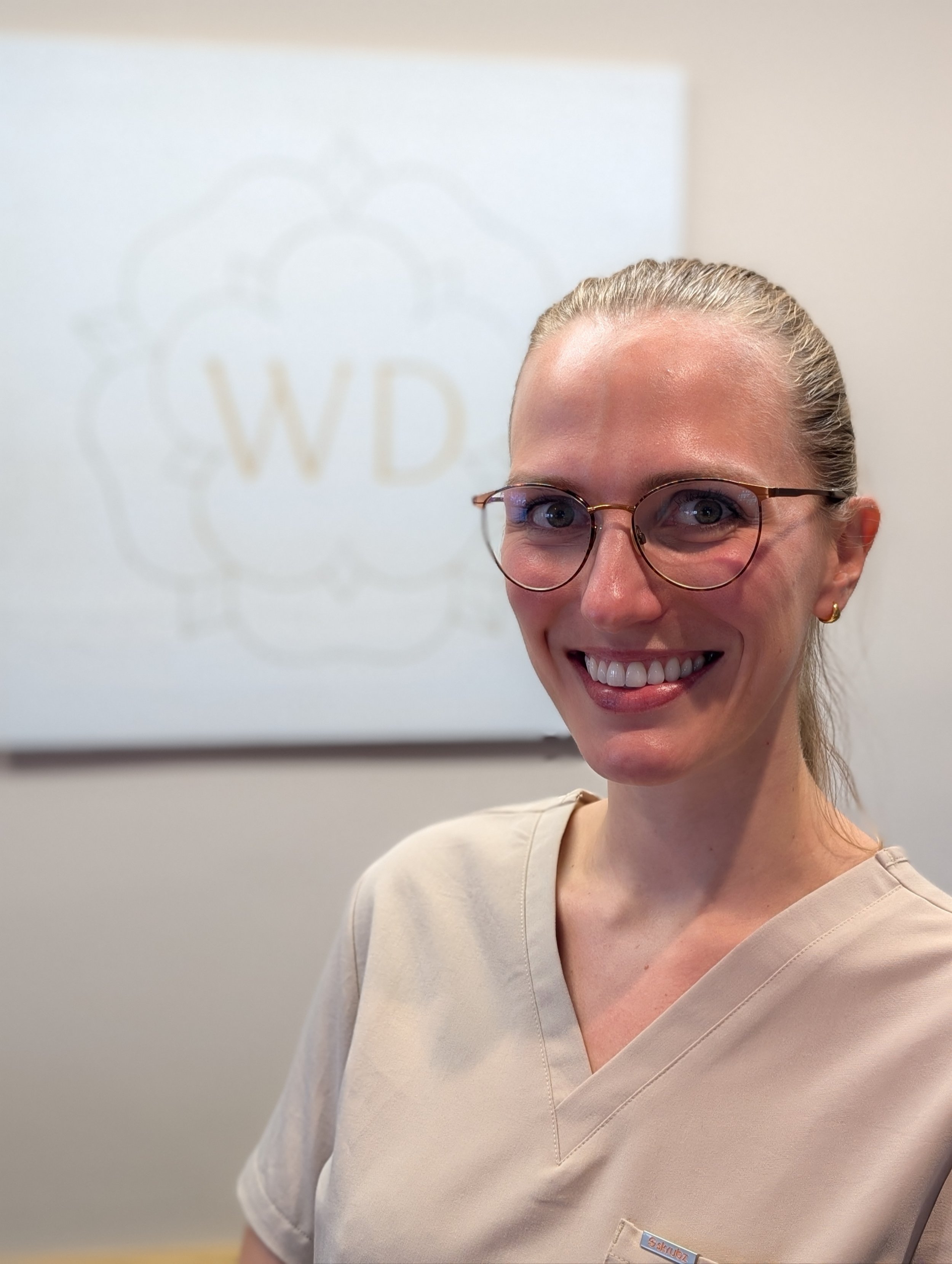 A smiling woman with blonde hair, wearing pink glasses, small hoop earrings, and a light green scrub top, standing against a plain beige background.