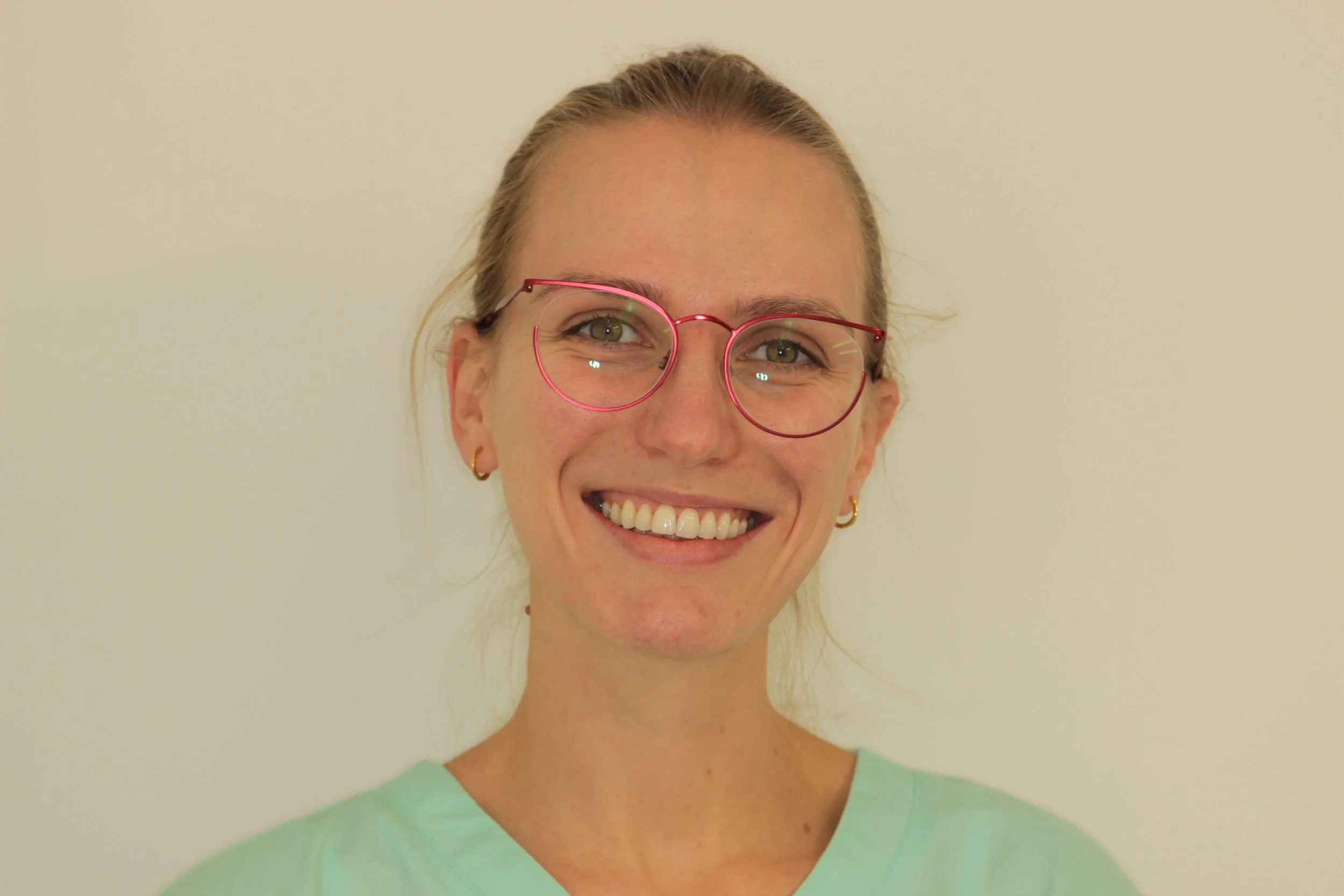 A smiling woman with blonde hair, wearing pink glasses, small hoop earrings, and a light green scrub top, standing against a plain beige background.