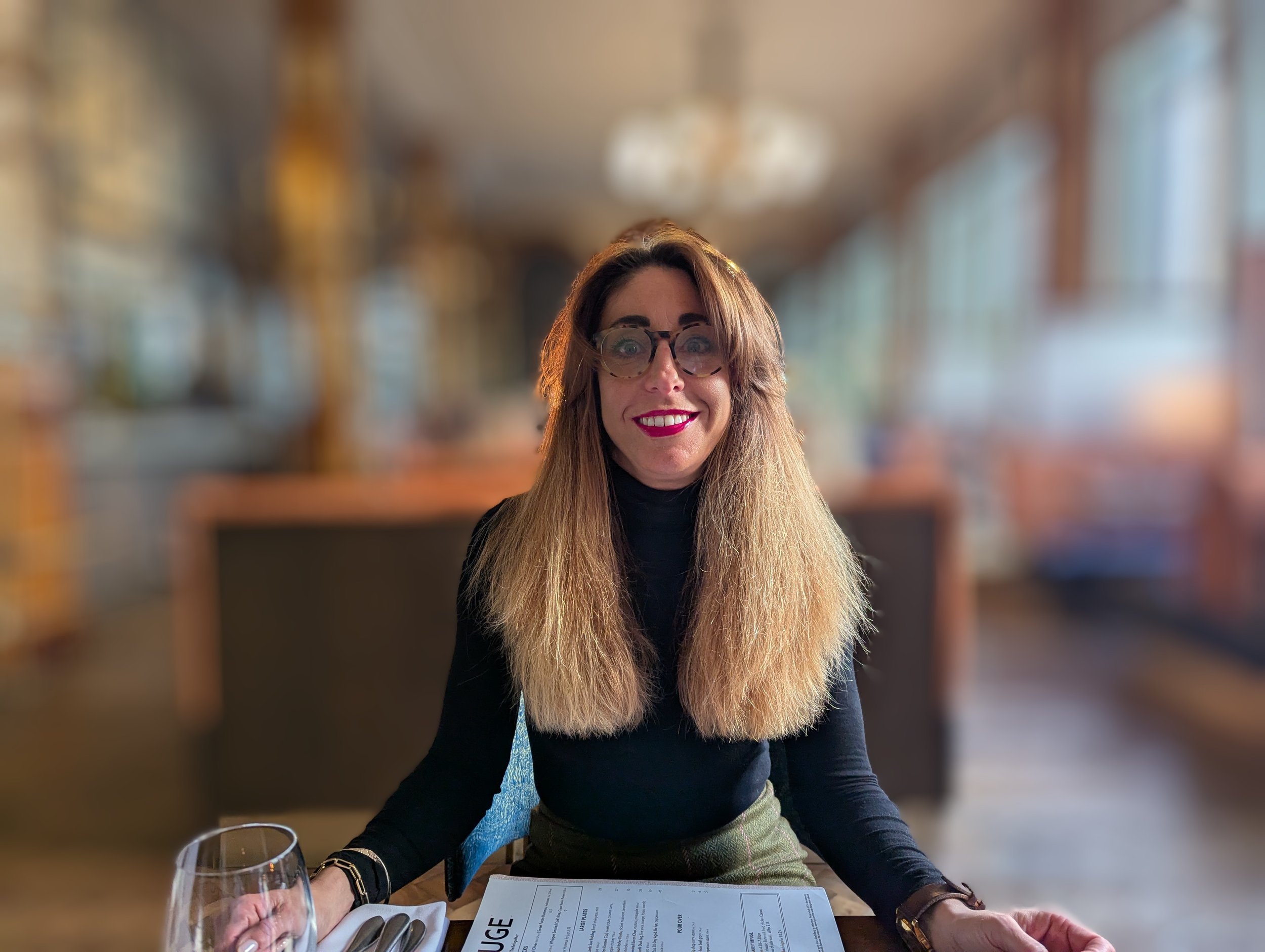 A woman with long, ombré hair, wearing glasses, red lipstick, a black turtleneck, seated at a table in a cozy restaurant or cafe, smiling at the camera with a menu, a glass of water, and silverware.