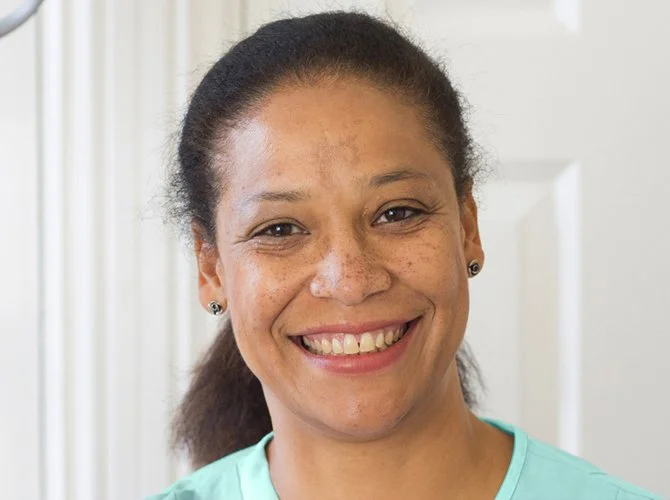 Close-up of a woman with dark hair pulled back, smiling, wearing a light blue top, standing indoors with a white door in the background.