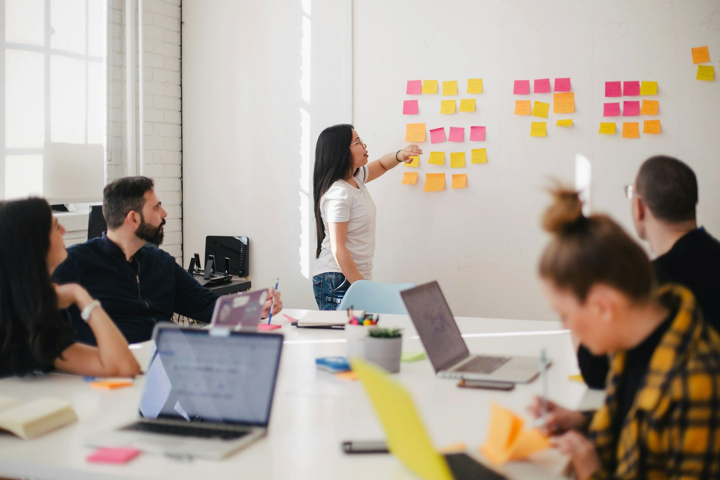 A group of people sit in a meeting, with laptops at a large table. One person stands at a whiteboard with a grid of colored sticky-notes