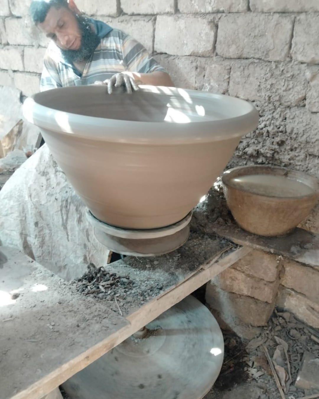 A man working on a large pottery wheel, shaping a large clay bowl in a rustic workshop with brick walls.