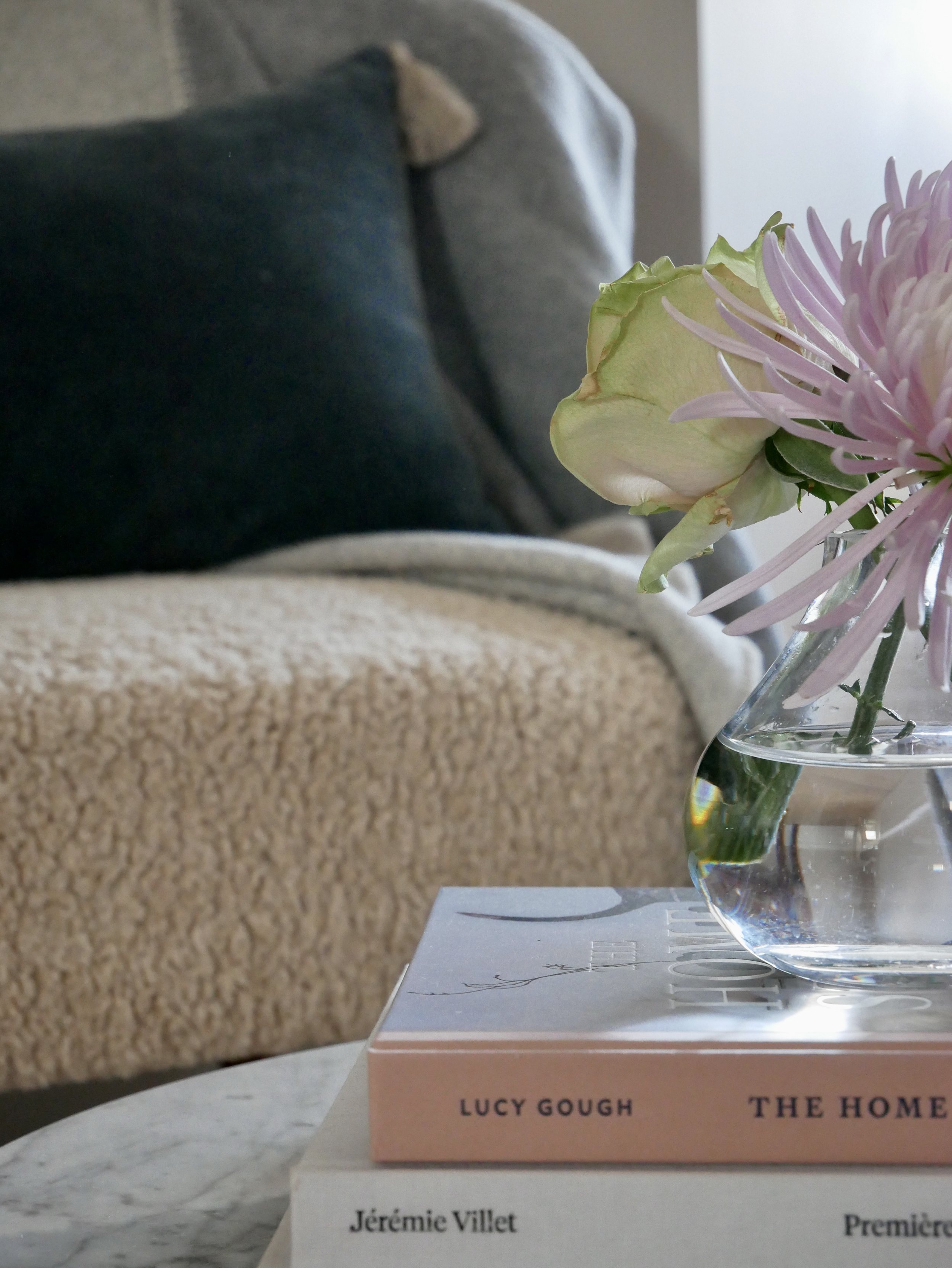 A close-up of a small glass vase with pink and white flowers placed on two stacked books on a table, with a textured beige couch and dark cushions in the background.